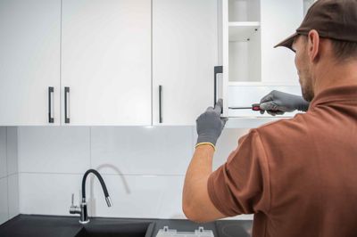 Technician installing a white kitchen cabinet with a drill in a modern kitchen