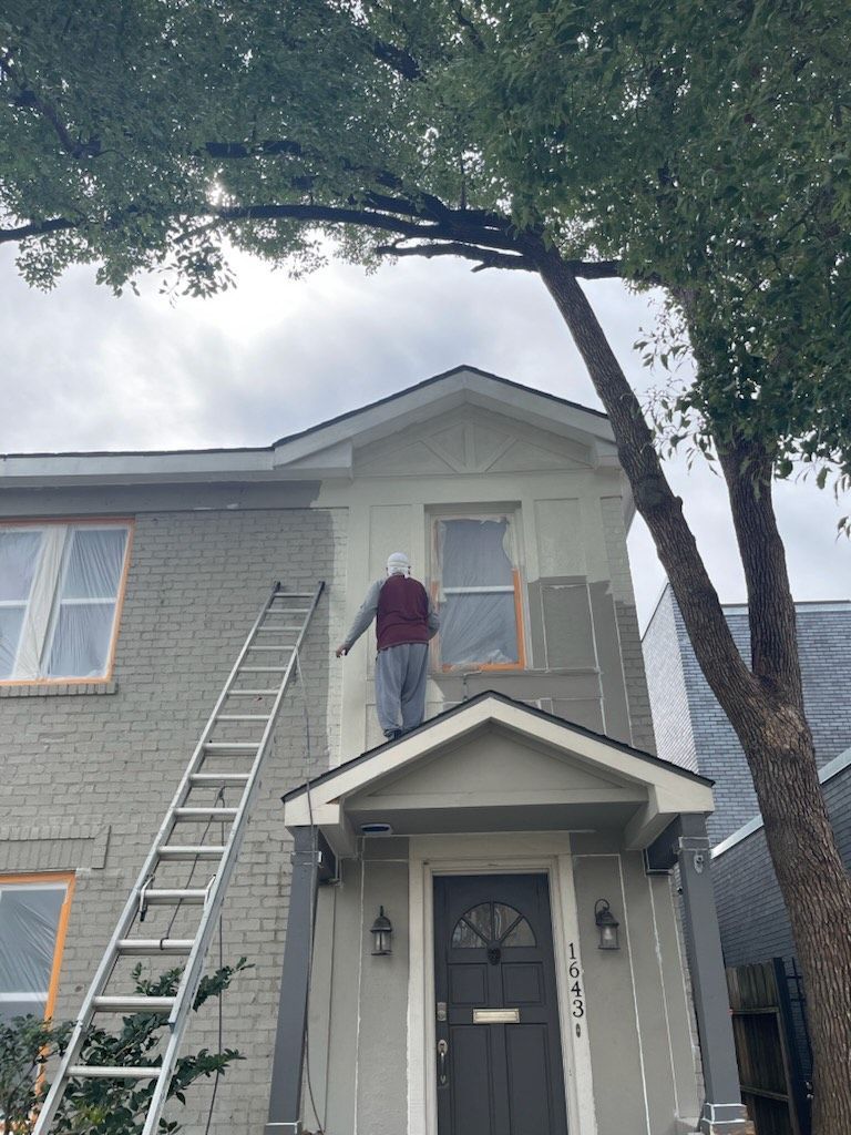 Person painting a black ceiling while standing on a wooden ladder, next to a large window.