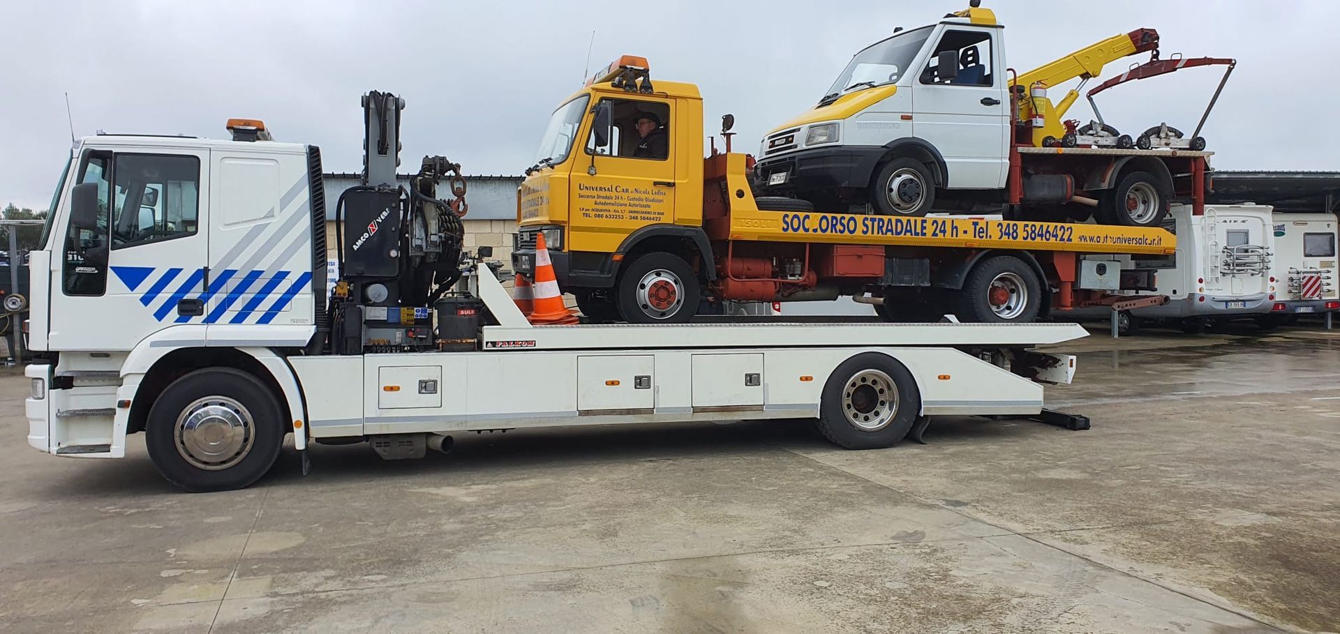 Un grande carro attrezzi bianco che trasporta un carro attrezzi giallo, il quale a sua volta trasporta un piccolo veicolo di traino bianco.