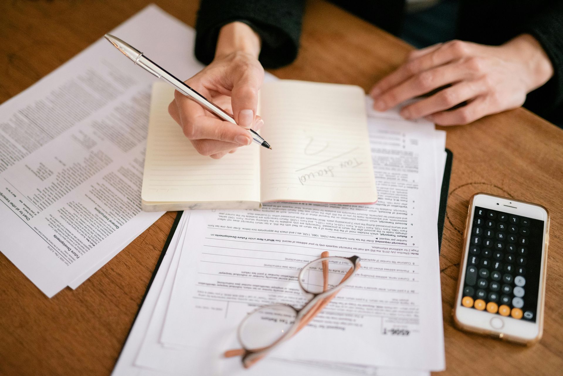 Person writing in a notebook on a desk with documents, a phone calculator, and glasses.