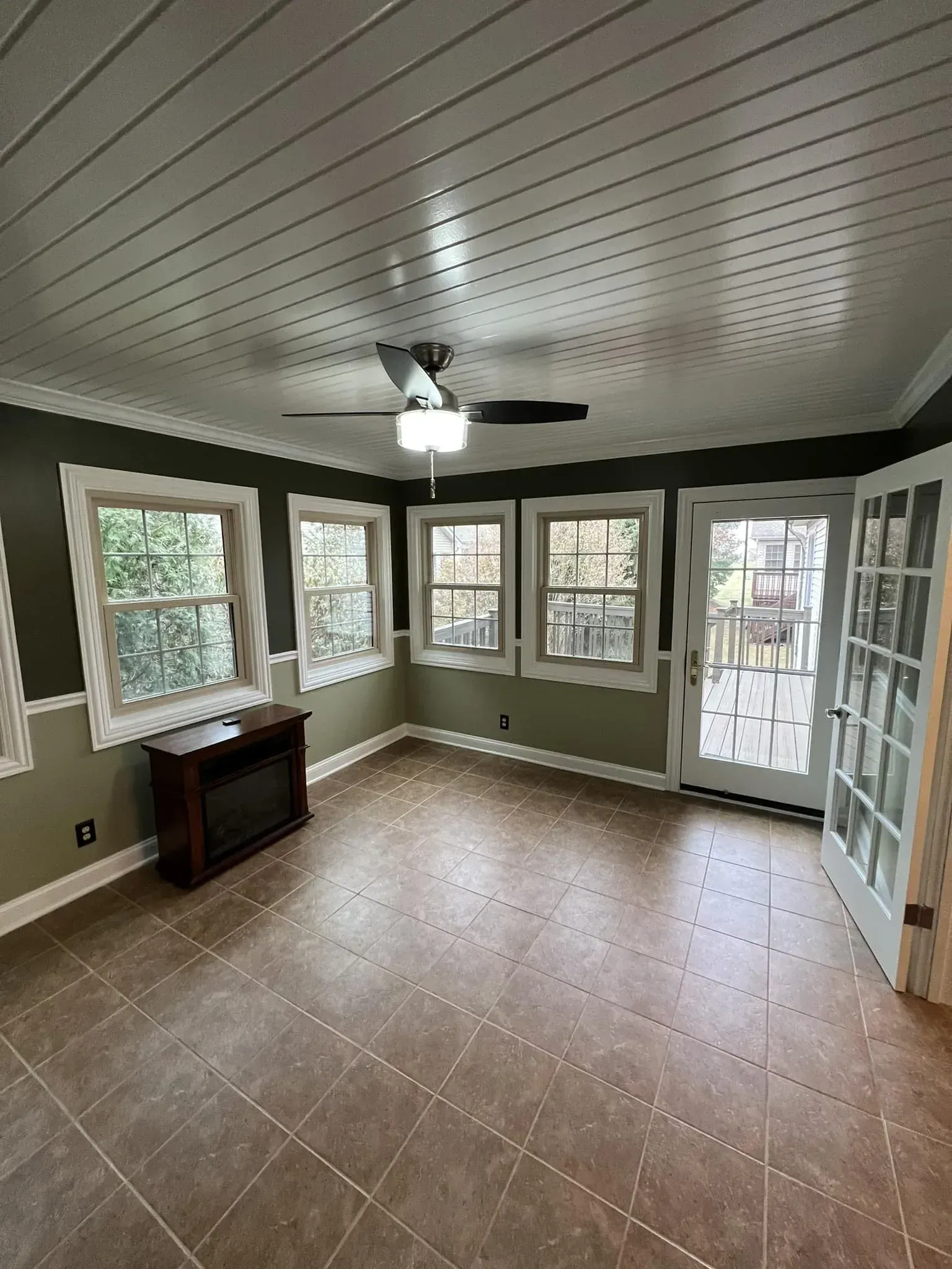 Empty sunroom with tan tile floor, white paneled ceiling, ceiling fan, and many windows with outdoor greenery.