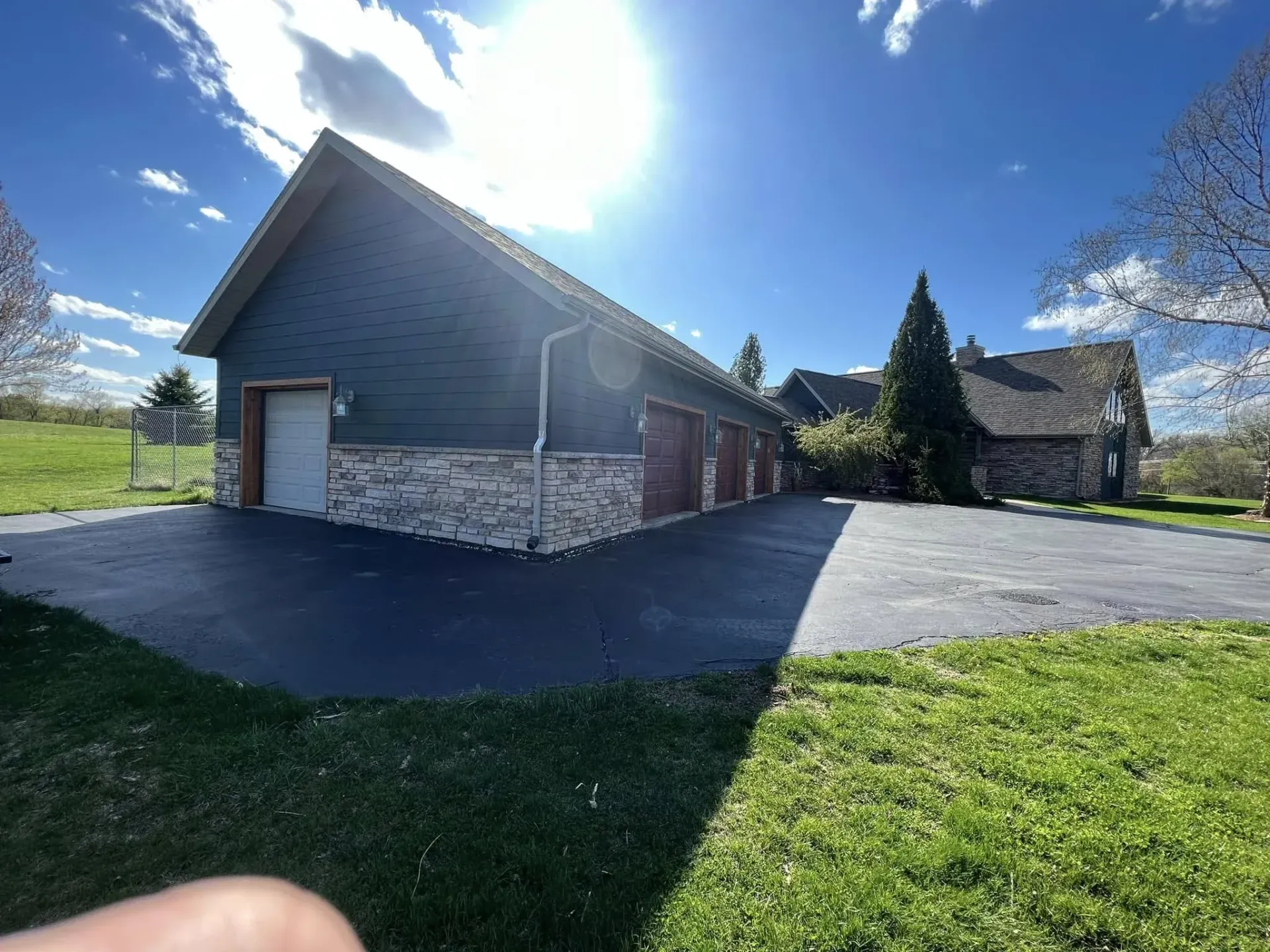 Wood-sided building with stone base, attached garage, and sunlit paved driveway under a blue sky