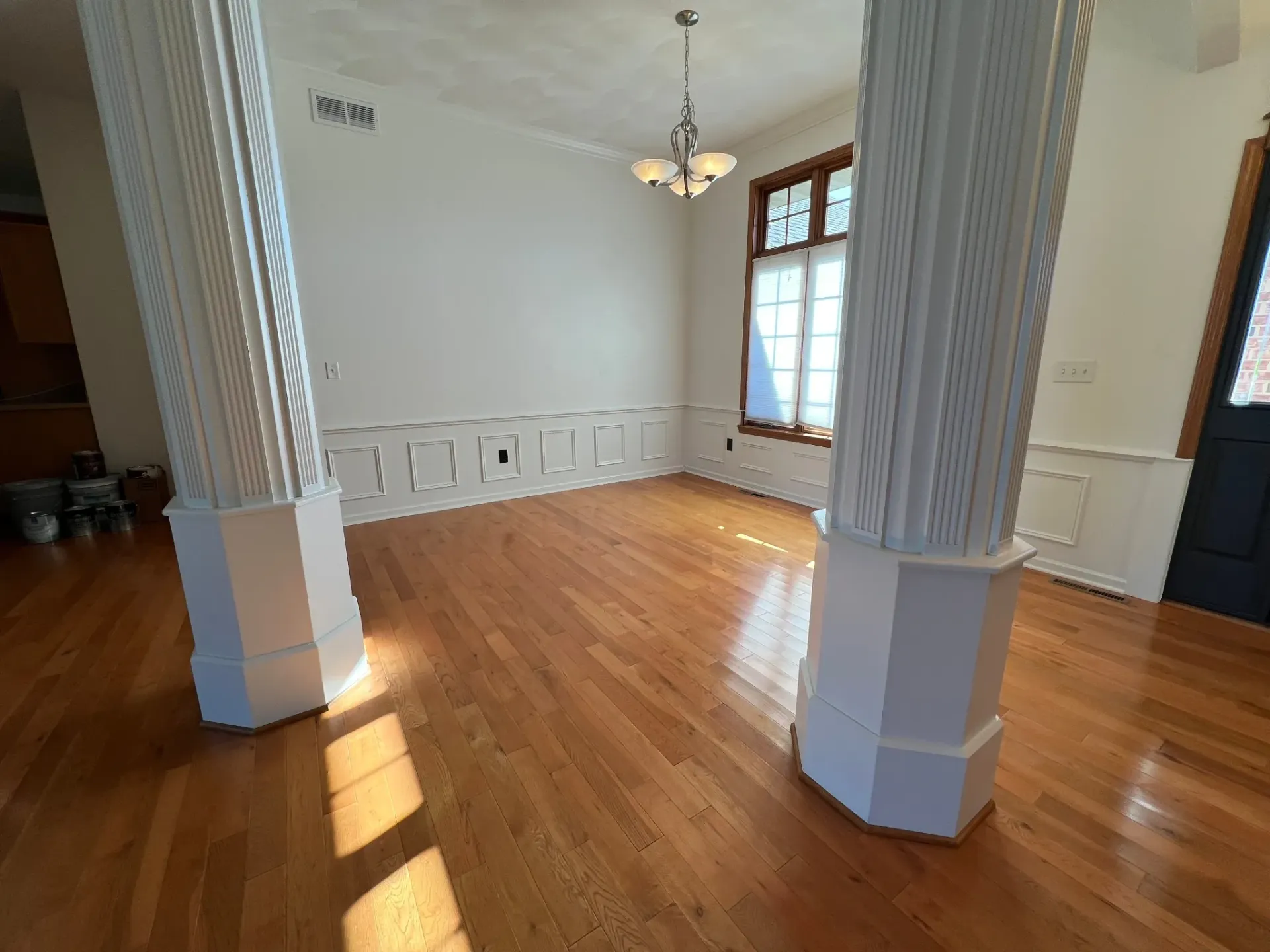 Empty room with hardwood floors, white walls, ornate columns, and sunlight by a window.