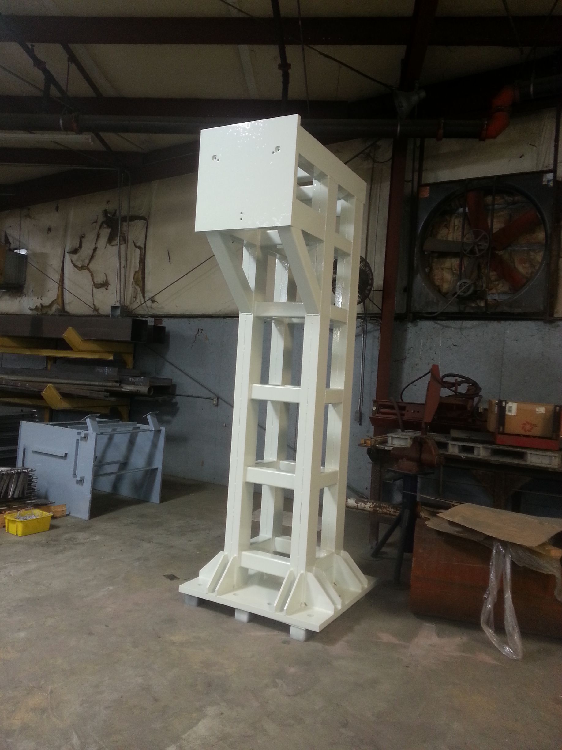 White, metal, tower-like structure with a square top and base, set in a workshop.