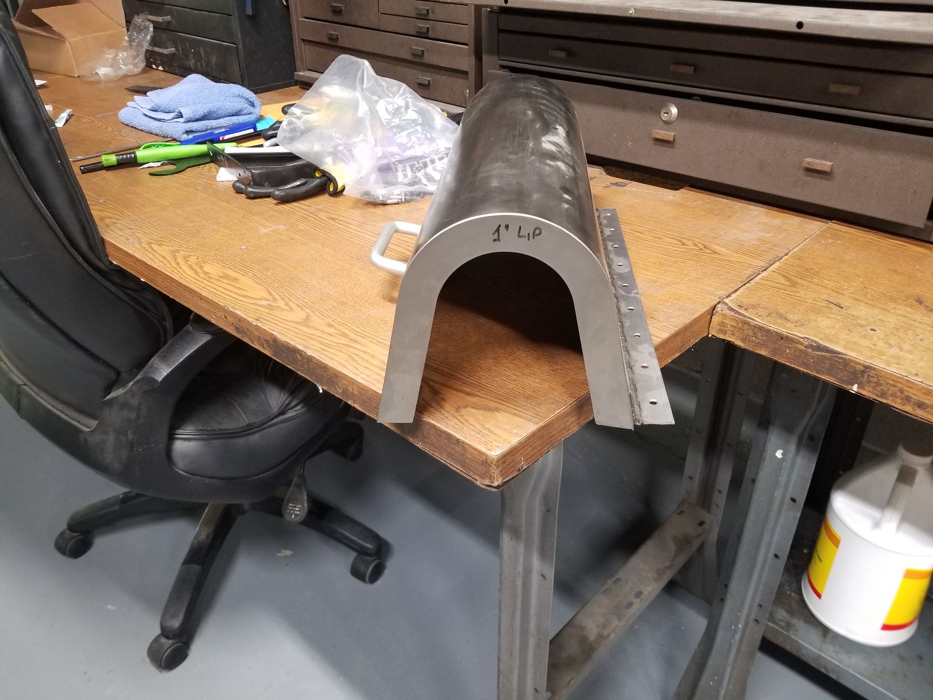 Metal arch-shaped component sits on a workbench with tools, beside a tool cabinet and a chair.