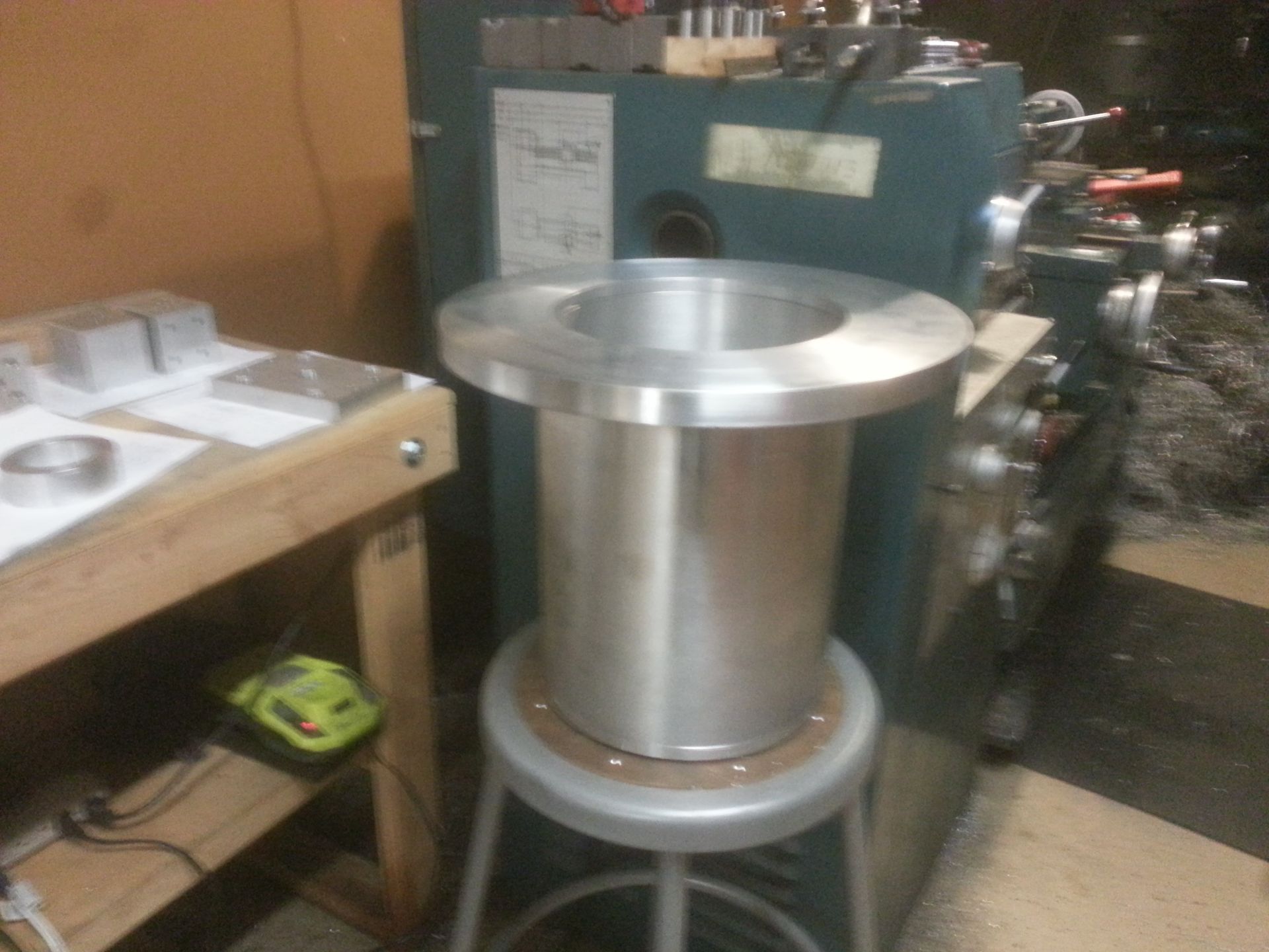 A large, cylindrical metal part with a wide flange on a stool, next to a metalworking lathe.