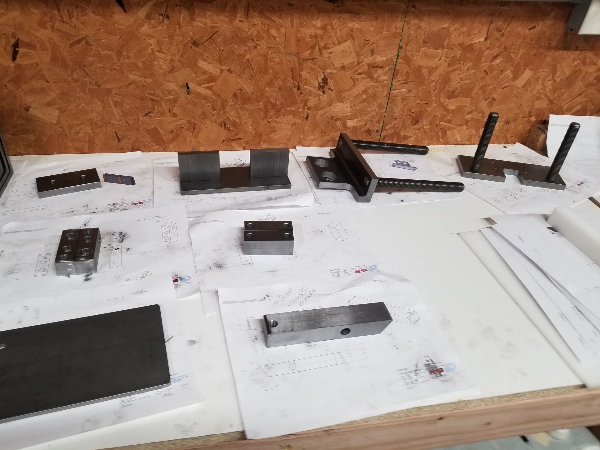 Metal parts, drawings, on a white covered workbench, against a wood background.