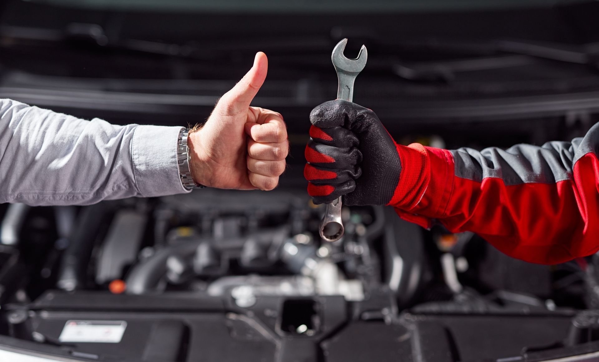 Two gloved hands exchange a thumbs-up and a wrench in front of a car engine.