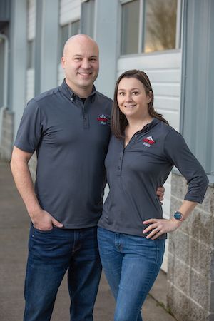 Two people in matching dark shirts stand smiling beside a building. | Ron's Auto & RV