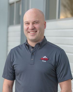 Smiling bald man in a gray polo shirt standing outdoors by a building | Ron's Auto & RV