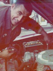 Man leaning into an open car engine bay, smiling while working on the engine. | Ron's Auto & RV
