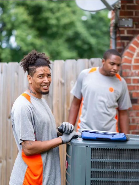 Two workers in gray shirts handling an outdoor HVAC unit beside a brick house and wooden fence.