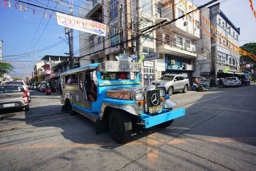 Colorfully decorated jeepney driving through a city street with buildings and parked cars in the background