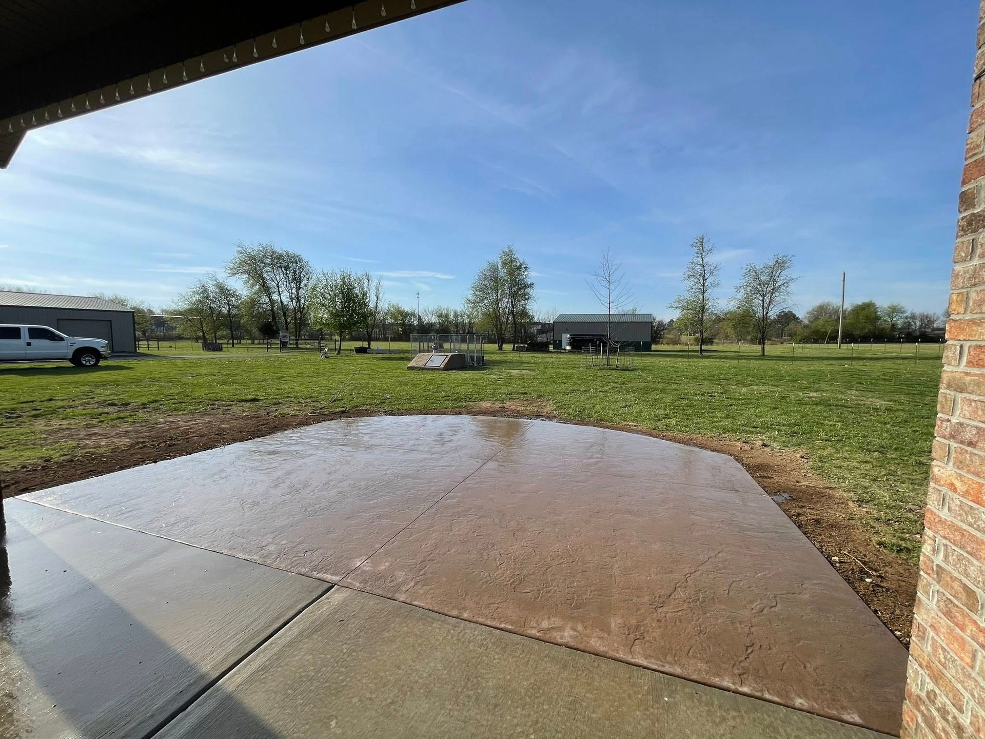 Wet concrete patio beside a grassy yard under a blue sky, with houses and trees in the distance