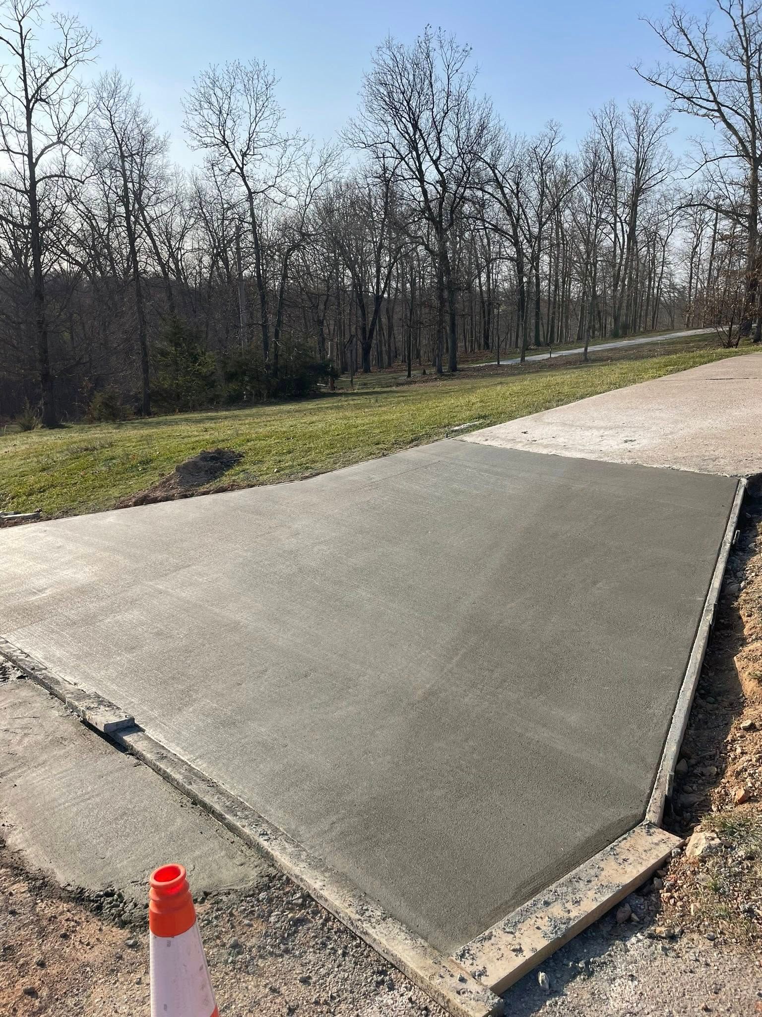 New concrete sidewalk ramp at the edge of a path, bordered by grass and leafless trees