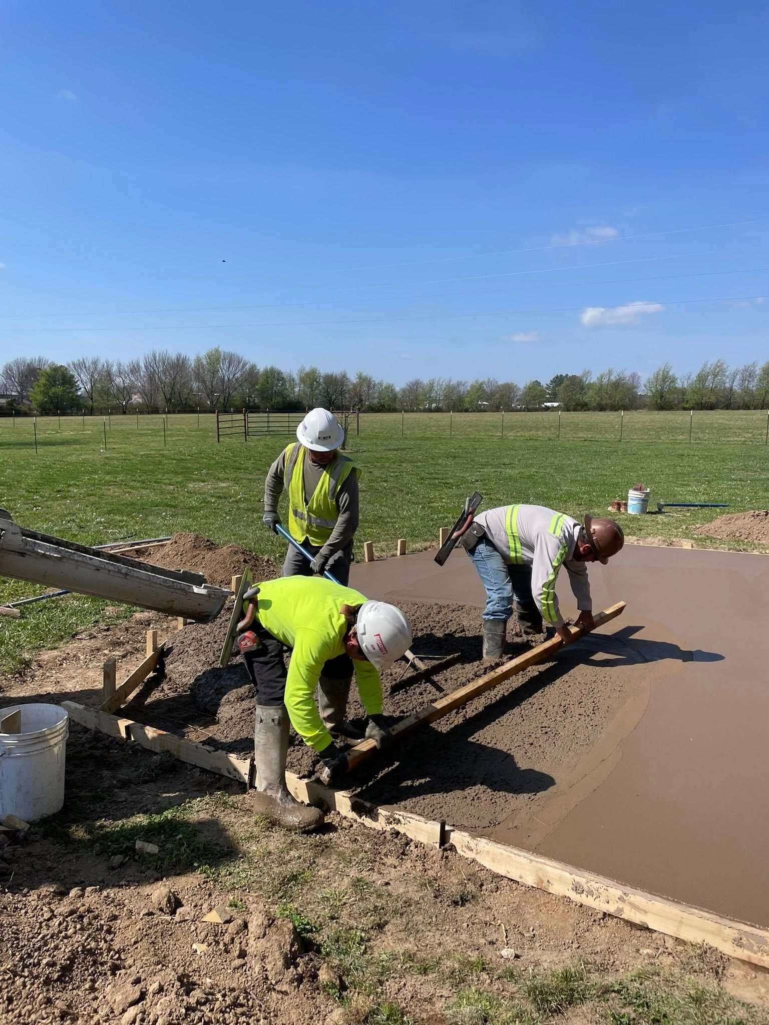 Workers smoothing fresh concrete beside a field under a blue sky
