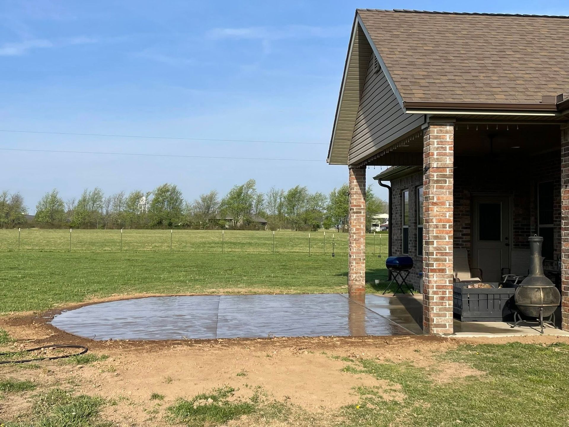 Backyard patio beside a brick house with a large puddle on the ground and open grassy field beyond
