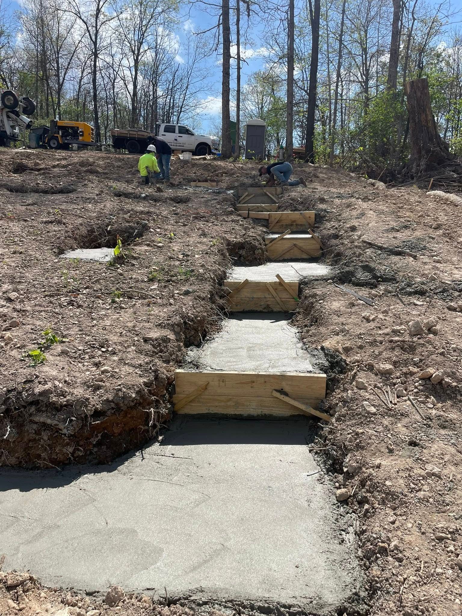 Concrete steps under construction on a wooded hillside, with workers and equipment in the background