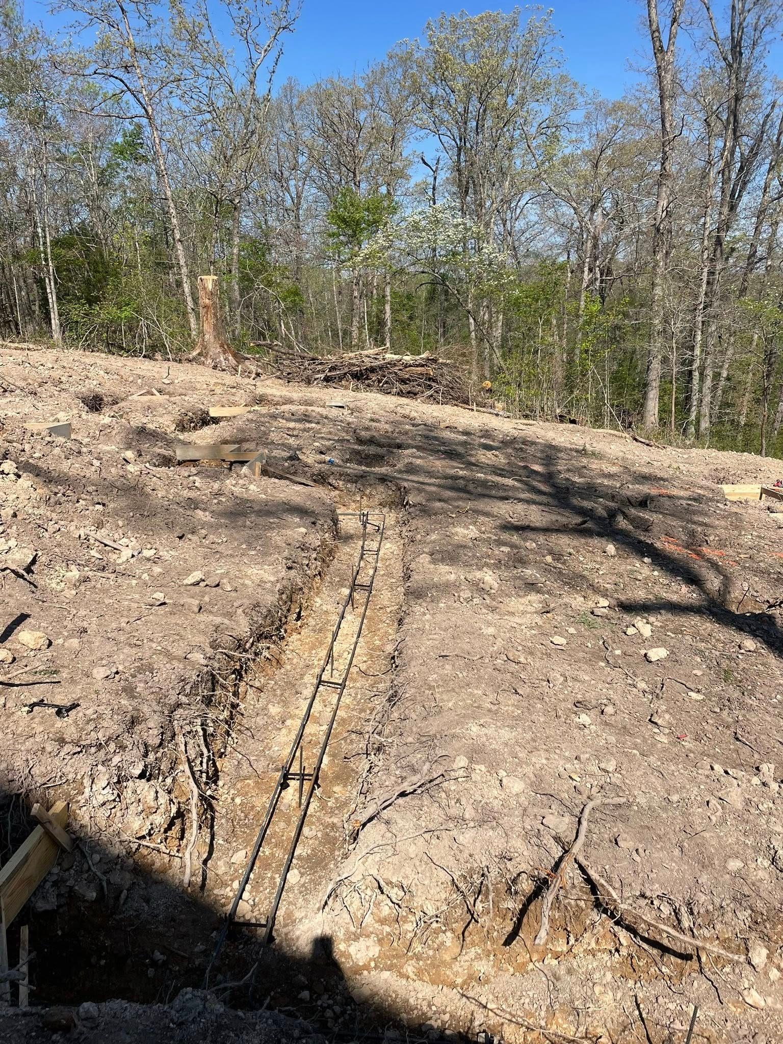 Cleared dirt hillside with a long trench and sparse trees under a bright blue sky