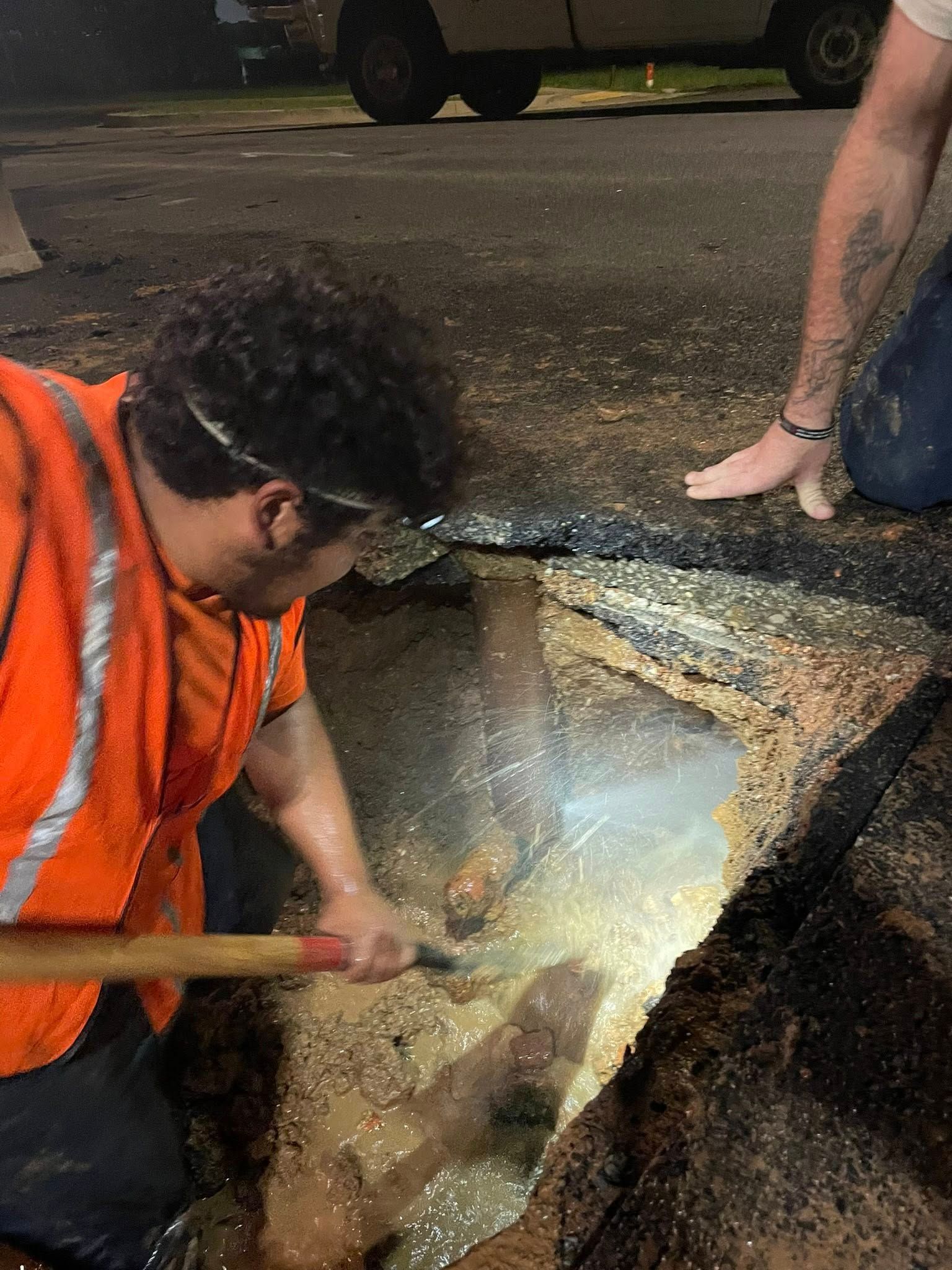 Worker in an orange vest examining a broken roadside hole with a flashlight at night