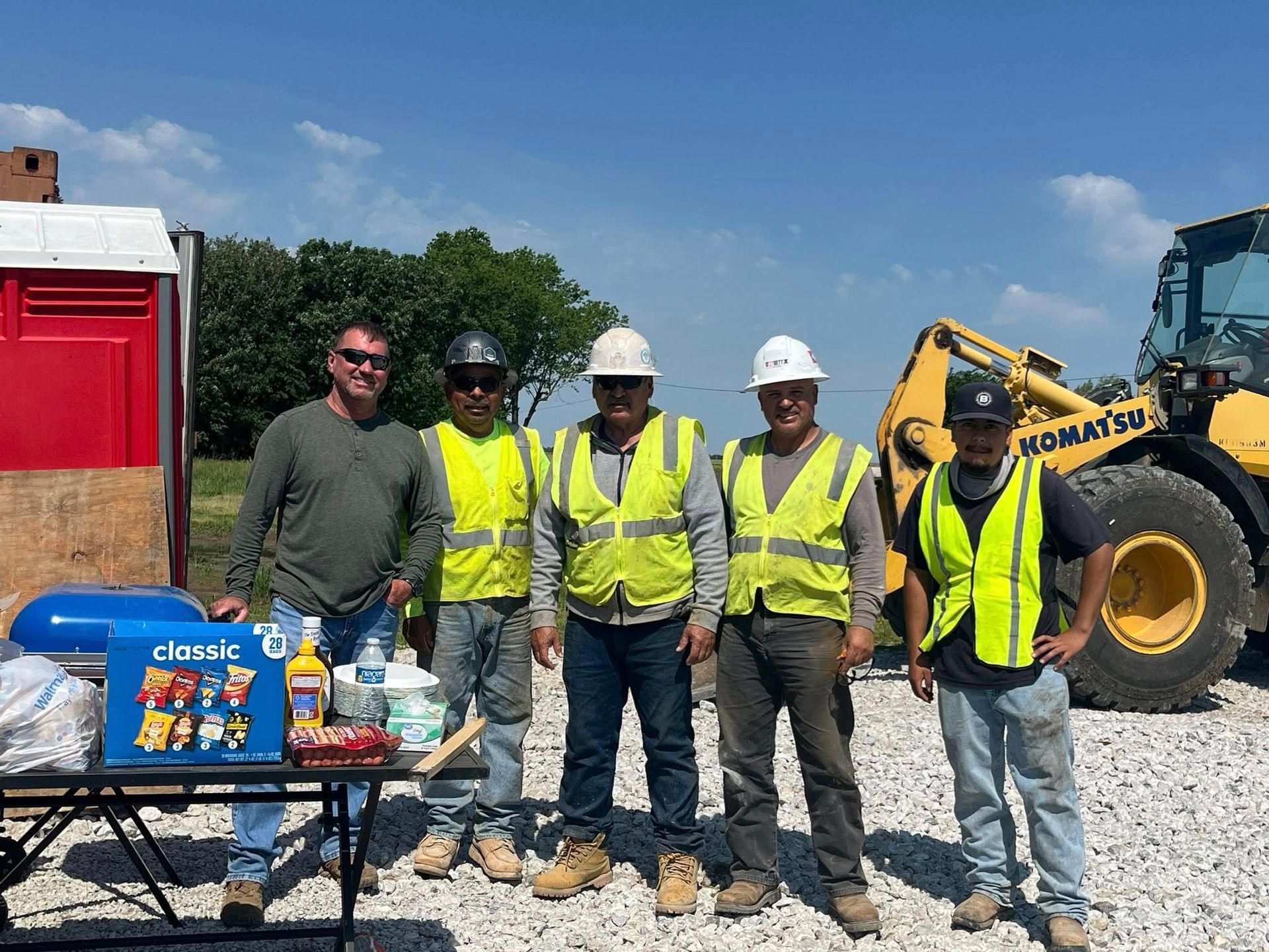 Five workers in neon safety vests and hard hats stand by a table of drinks and tools on a construction site.
