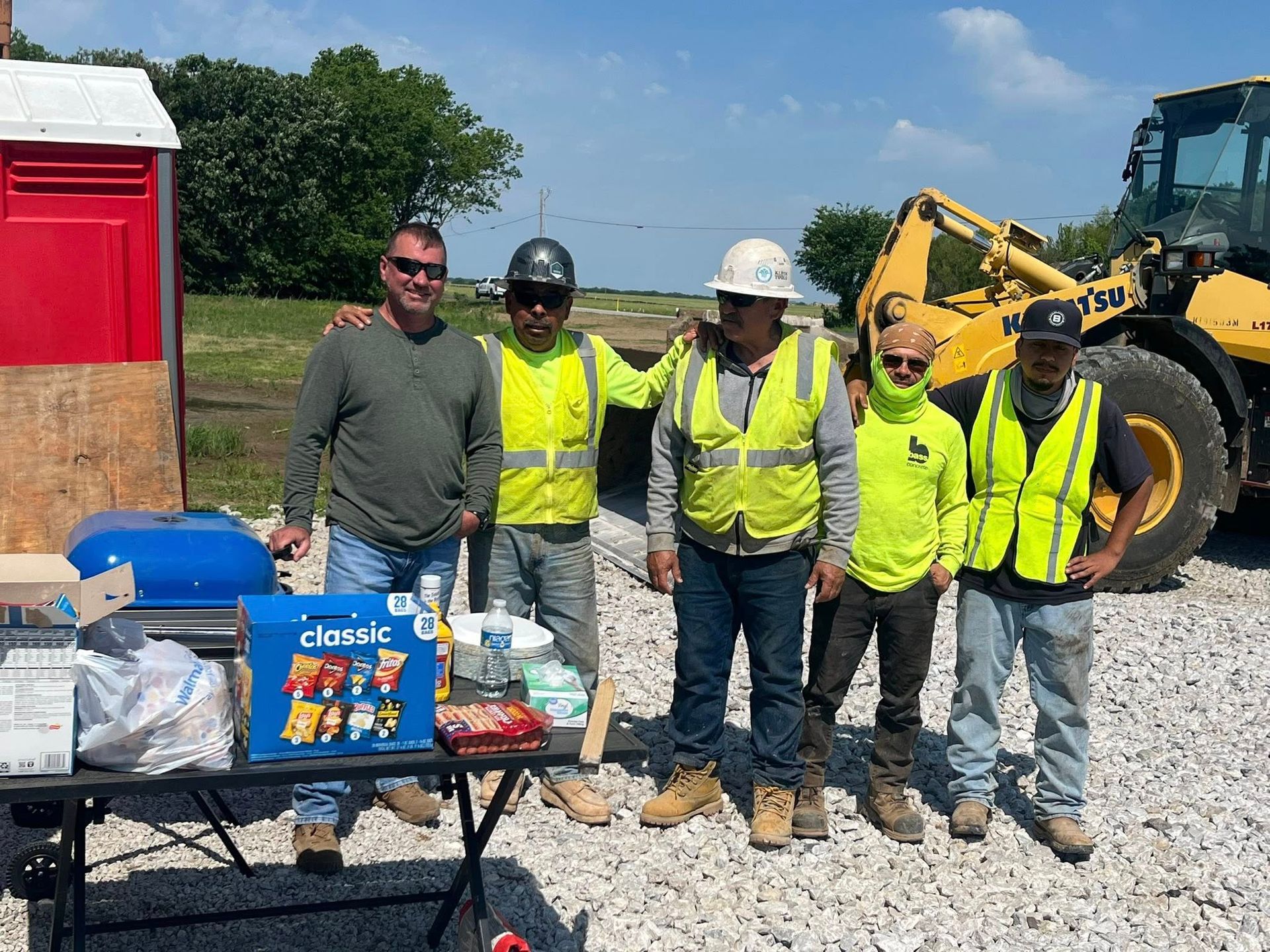 Five workers in safety vests and hard hats pose at a roadside worksite beside a table of supplies.