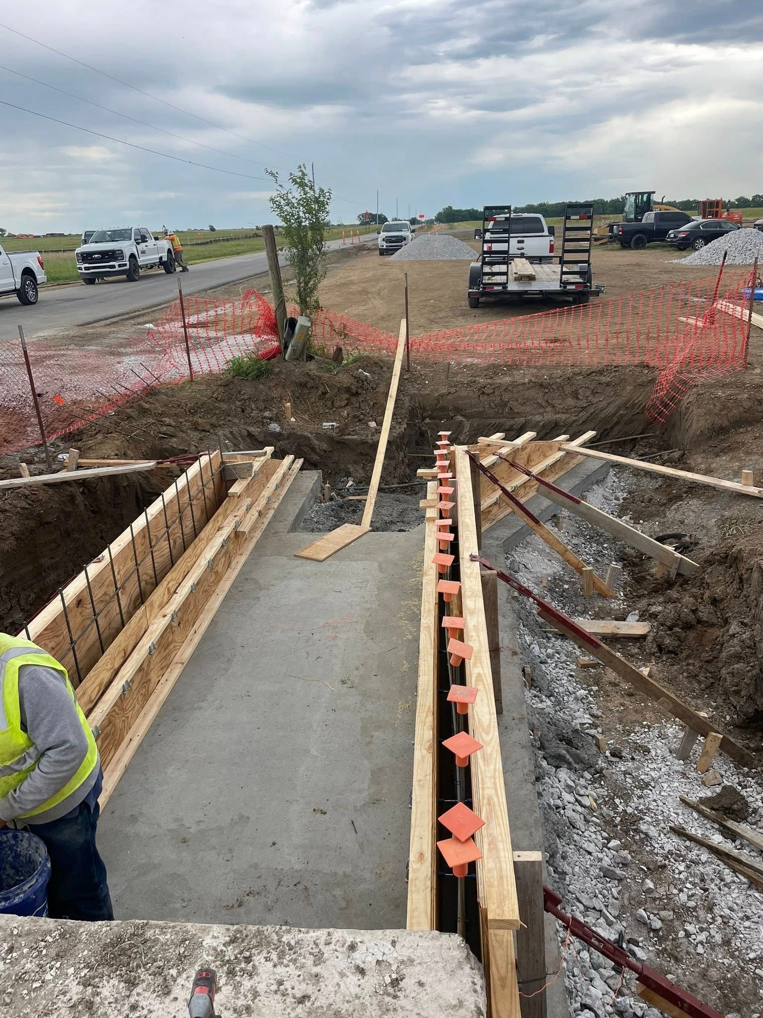 Road construction site with concrete forms, trench, excavator, and worker in a high-visibility vest