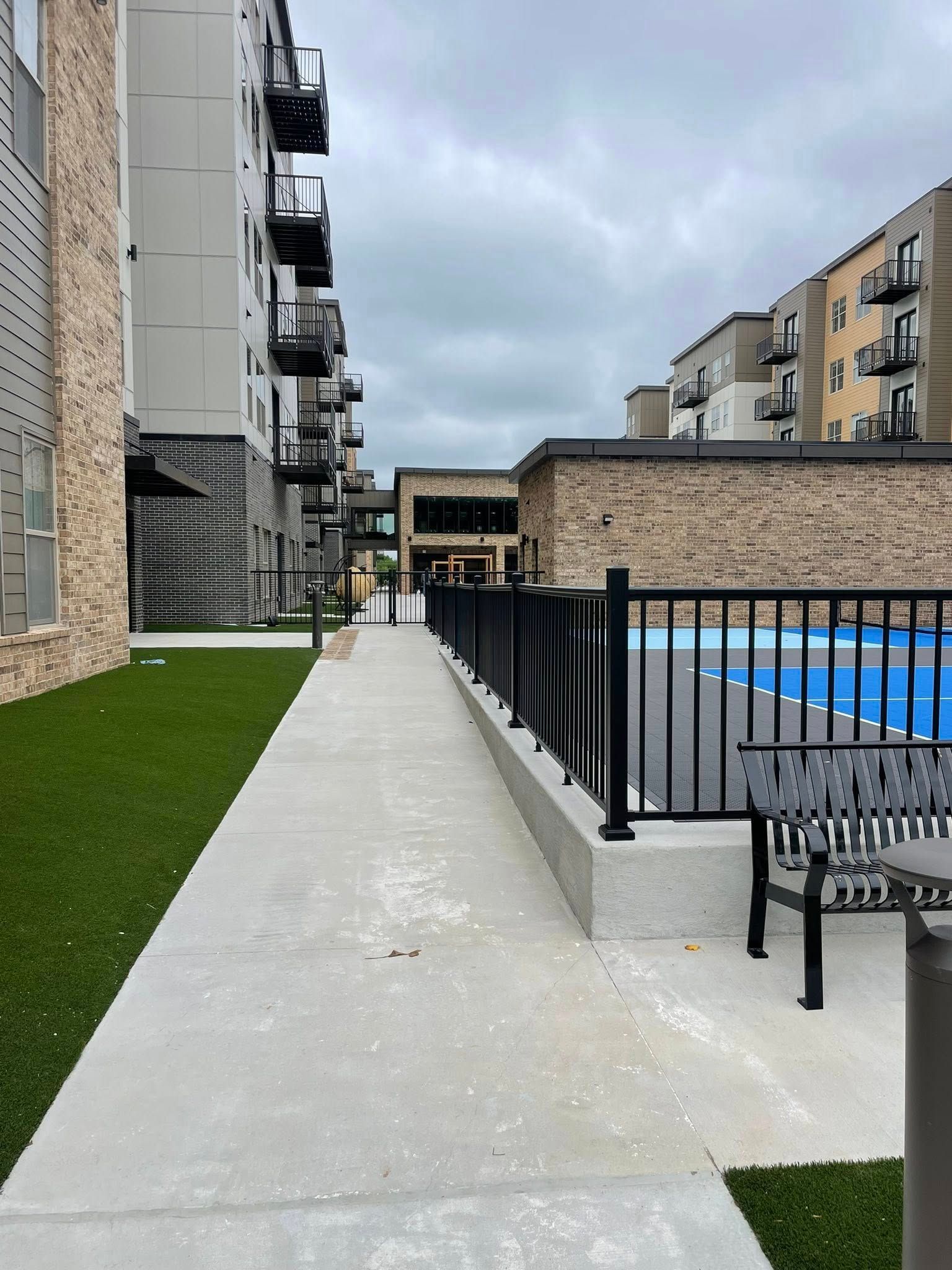 Apartment courtyard walkway beside a fenced blue pool, with benches and overcast sky.