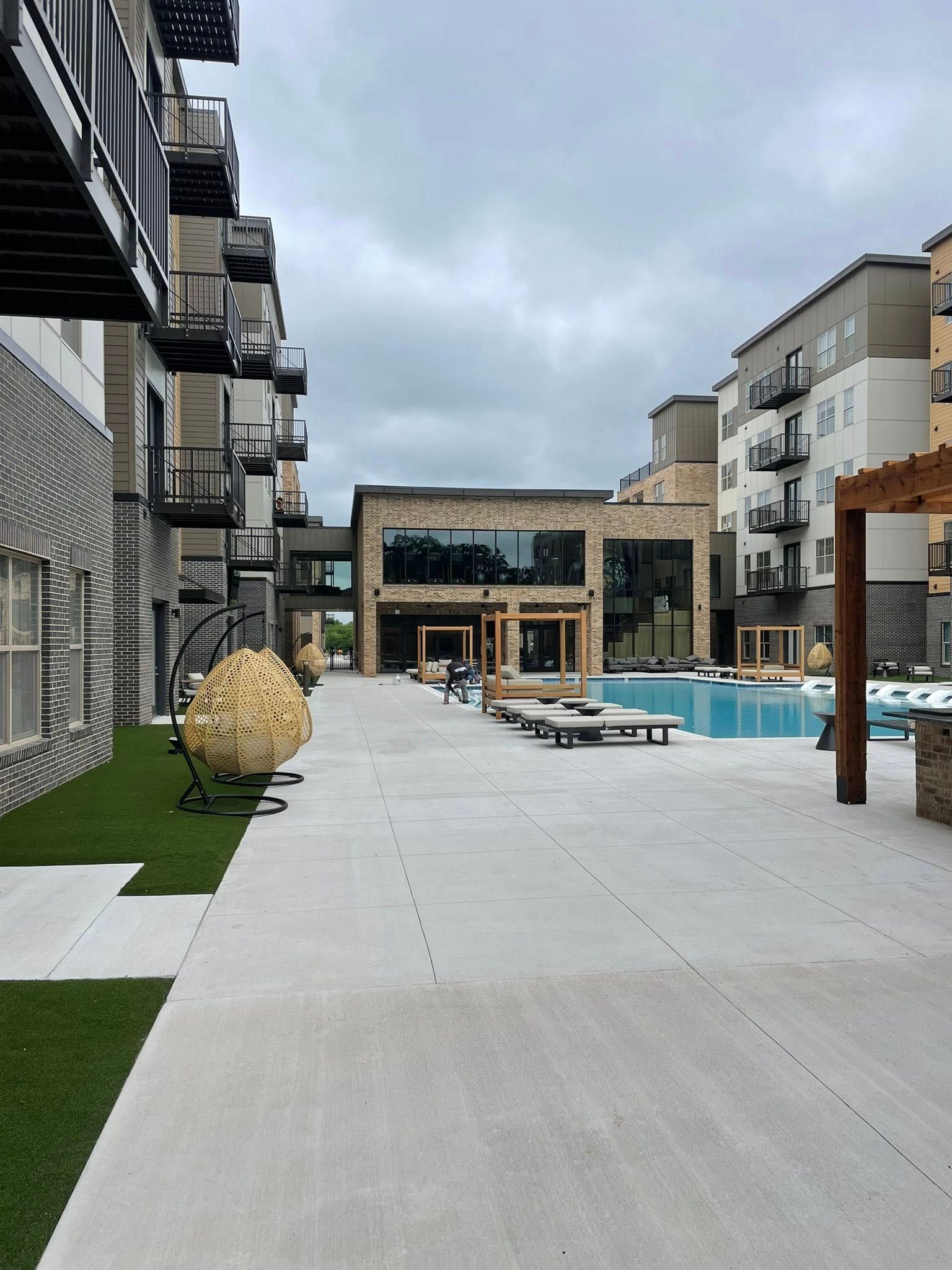 Modern apartment courtyard with pool, lounge chairs, and gray paved walkway under cloudy sky