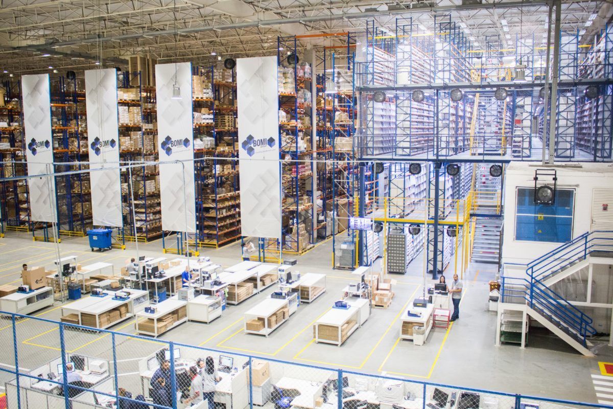 Warehouse interior with multi-level shelving, workers at packing stations, and overhead systems.