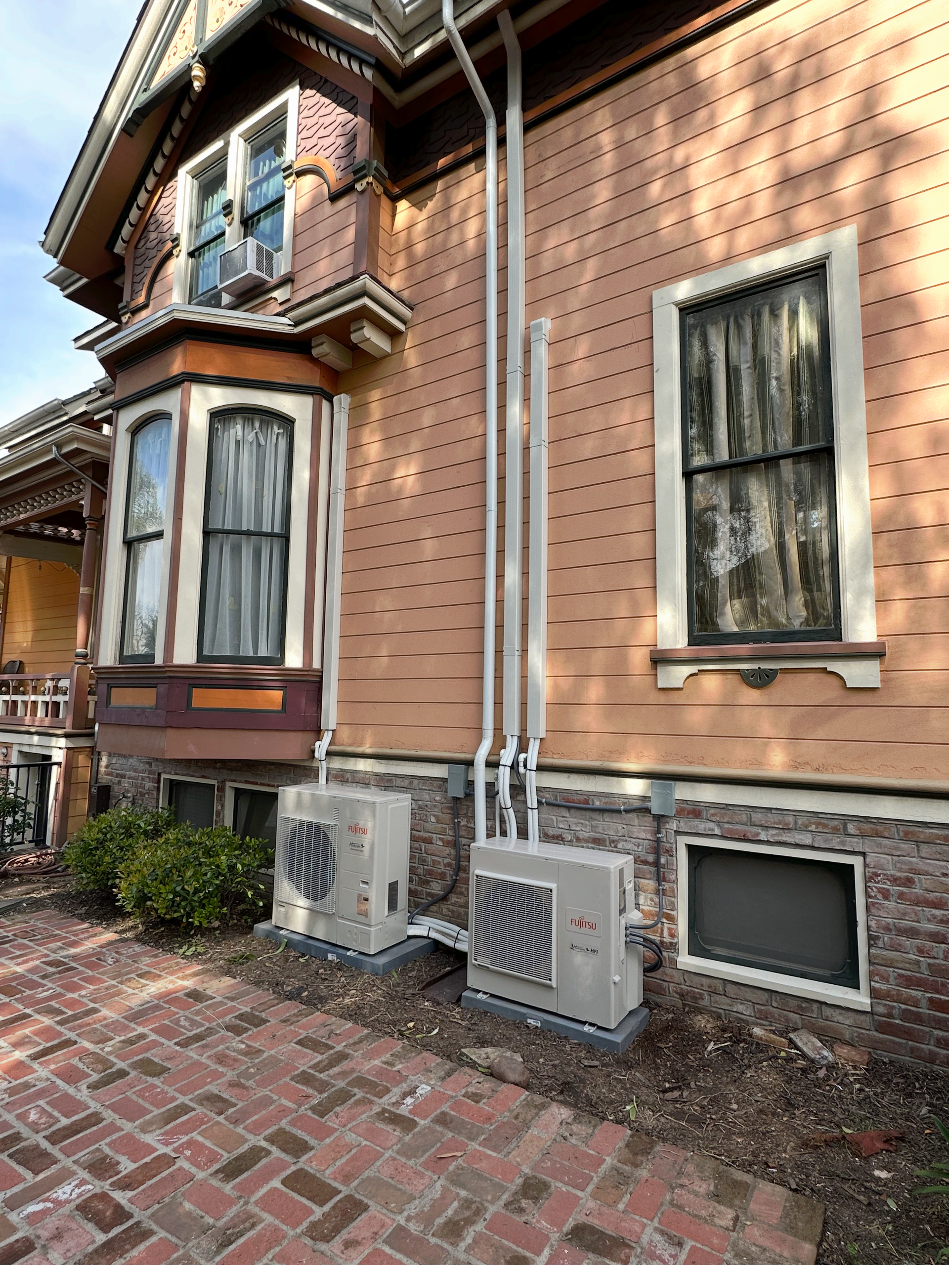 Two HVAC units mounted on an old brick building. White pipes run up the building's side.