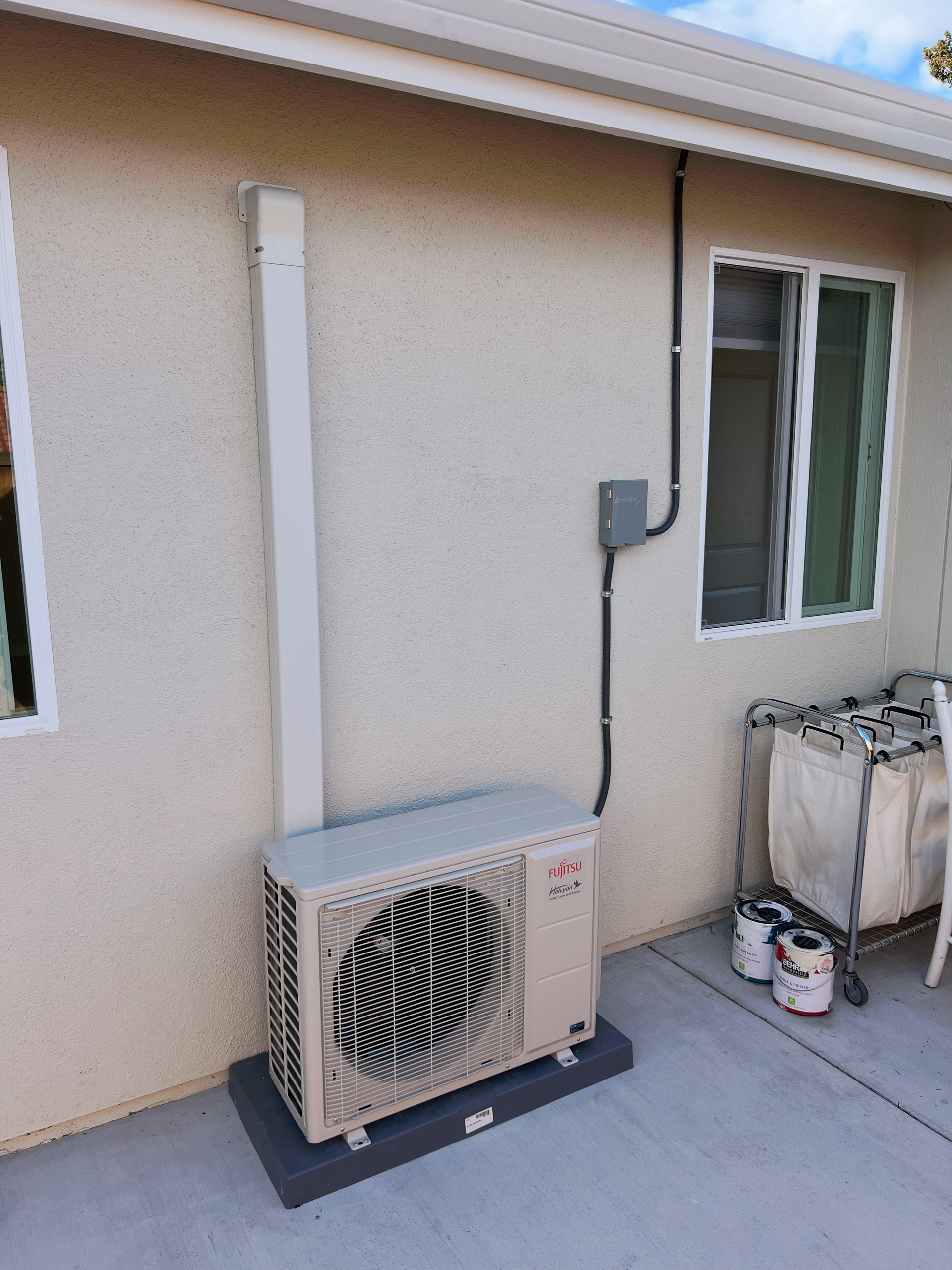 Outdoor air conditioning unit mounted on a gray pad against a stucco wall. Electrical conduit and box present.