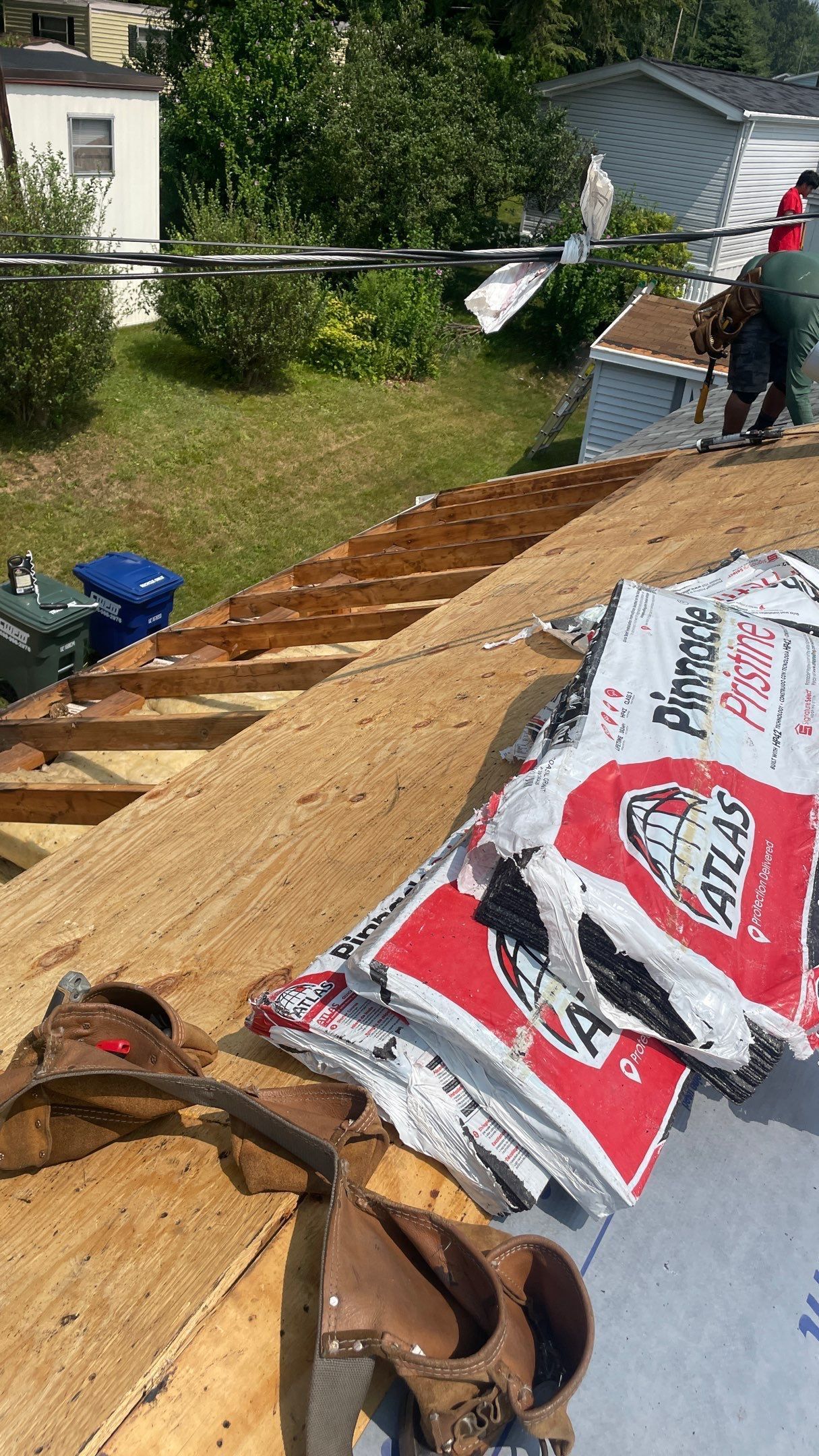 A high-angle view shows a roof undergoing construction with exposed wood, roofing felt, and stacked bundles of shingles.