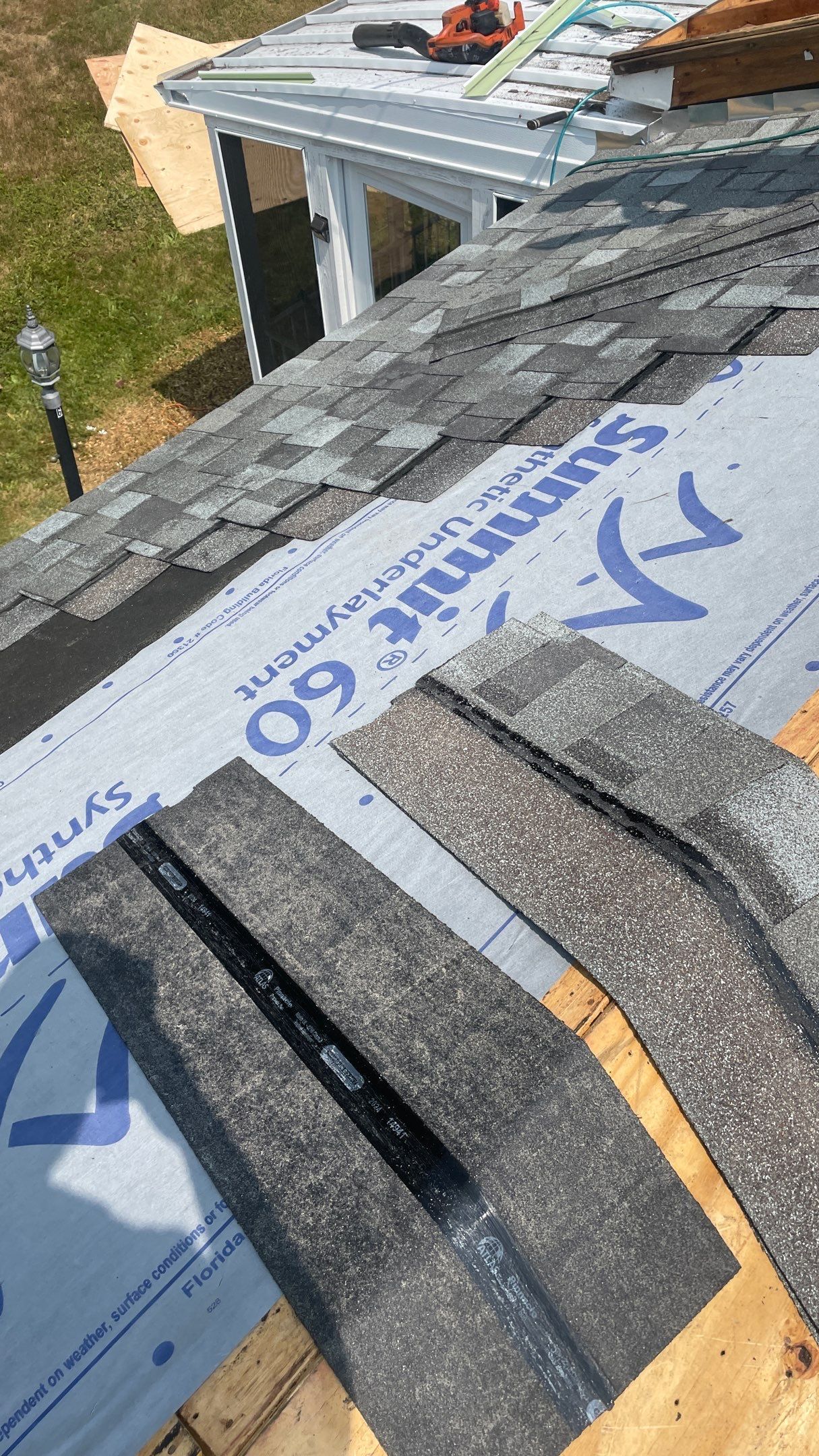 A close-up view of asphalt shingles being installed over gray synthetic roof underlayment on a wooden roof deck.