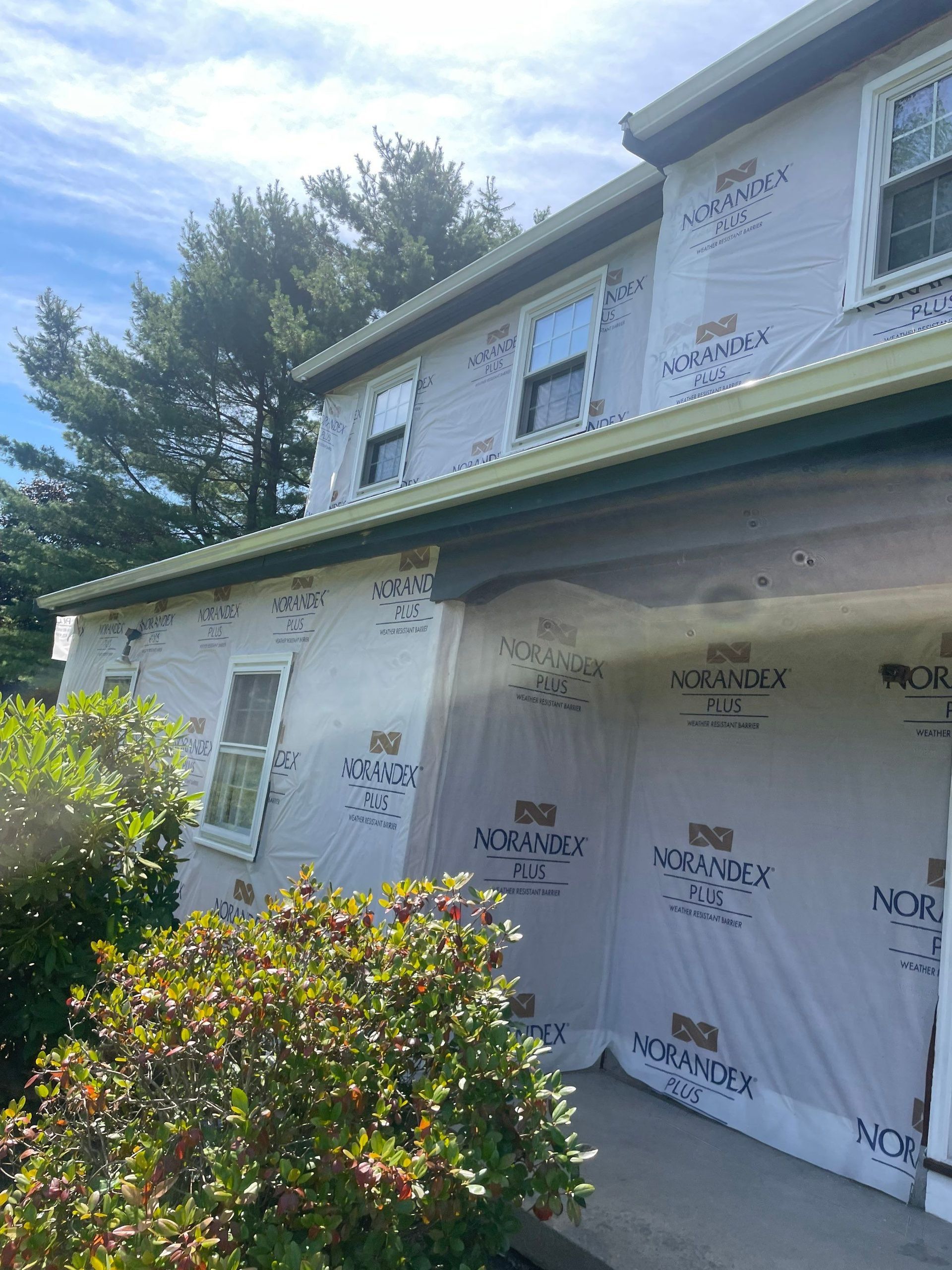 A house under construction with white house wrap, dark window frames, and green trim on a sunny day.