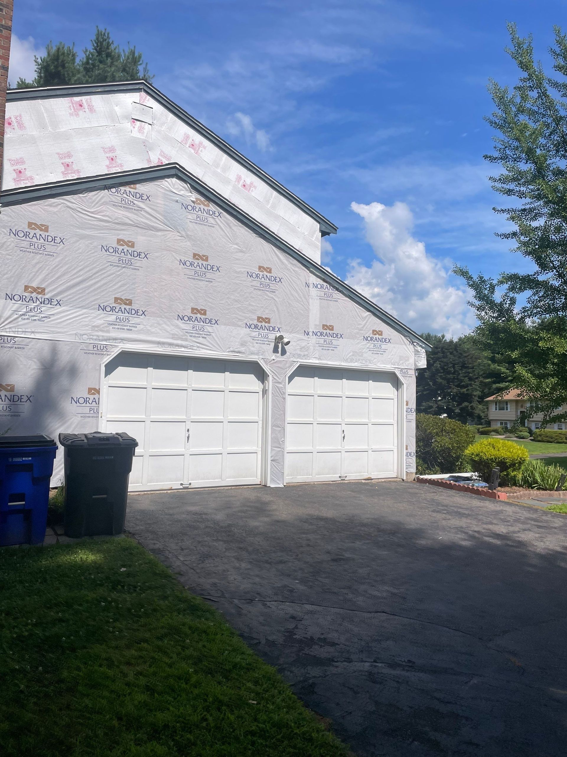 A house exterior under construction with white protective house wrap covering the walls and two white garage doors.