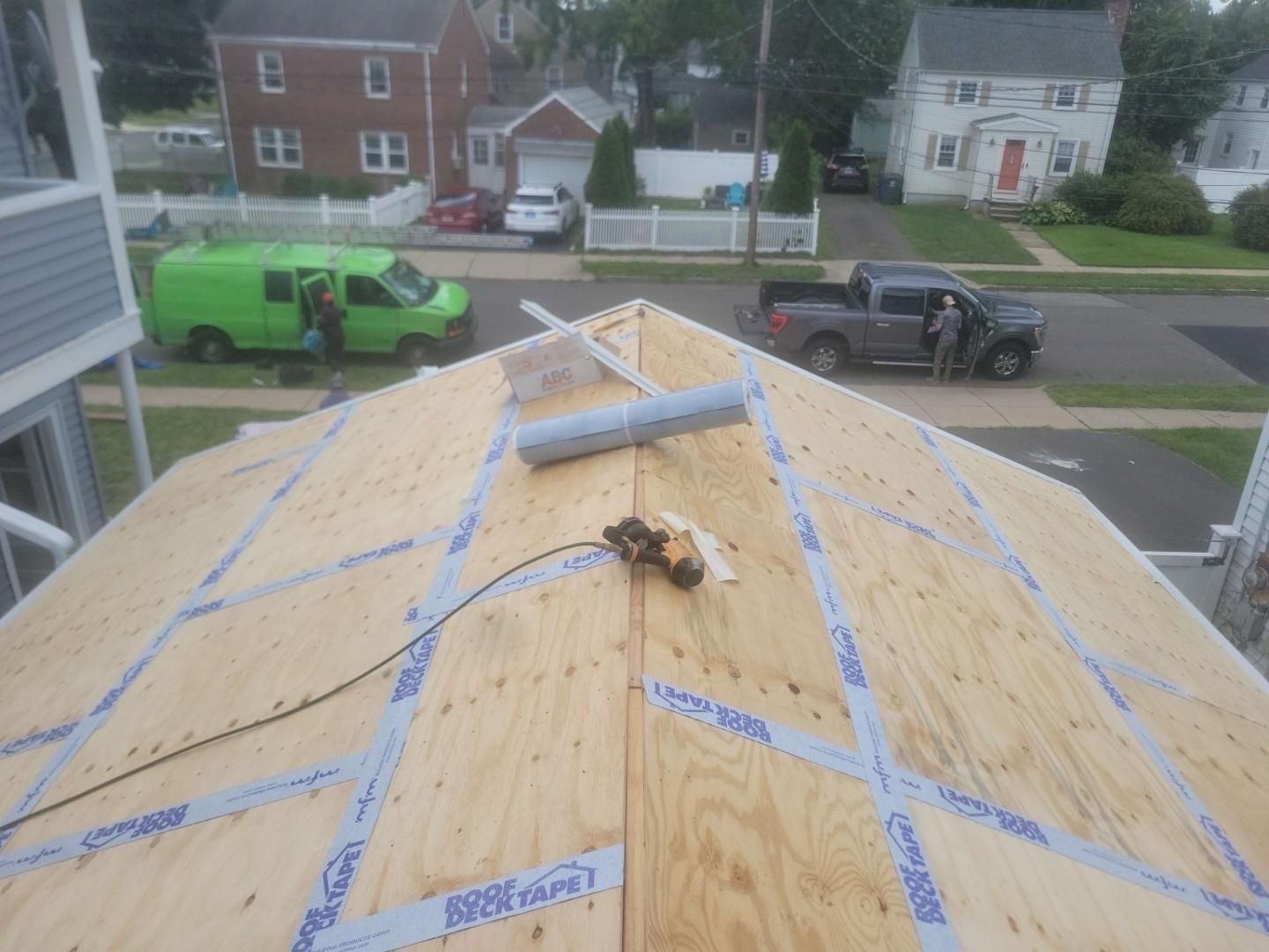 View from a roof under construction with plywood decking, seam tape, and tools, overlooking a suburban street.