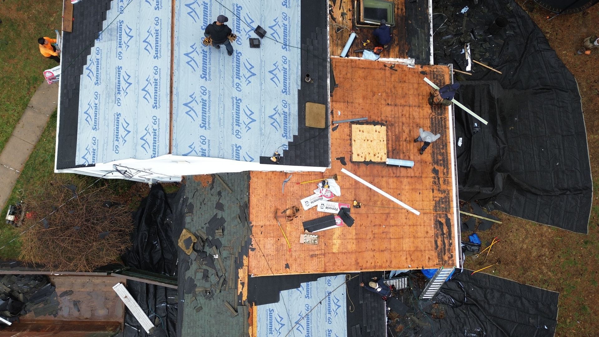 An aerial view of a residential roof under construction, featuring workers, exposed wood sheathing, and blue underlayment.