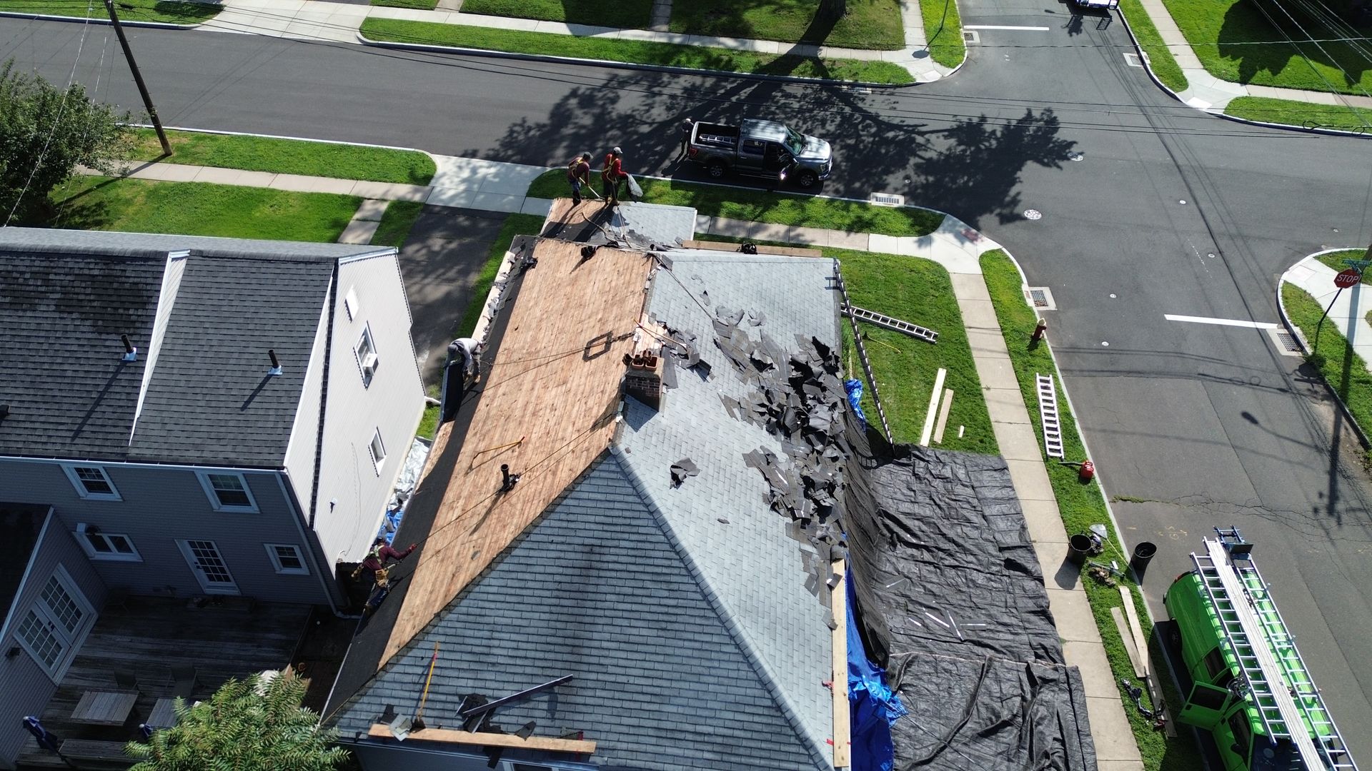 An aerial view of workers repairing a house roof, showing exposed wood decking, old shingles, and construction equipment.