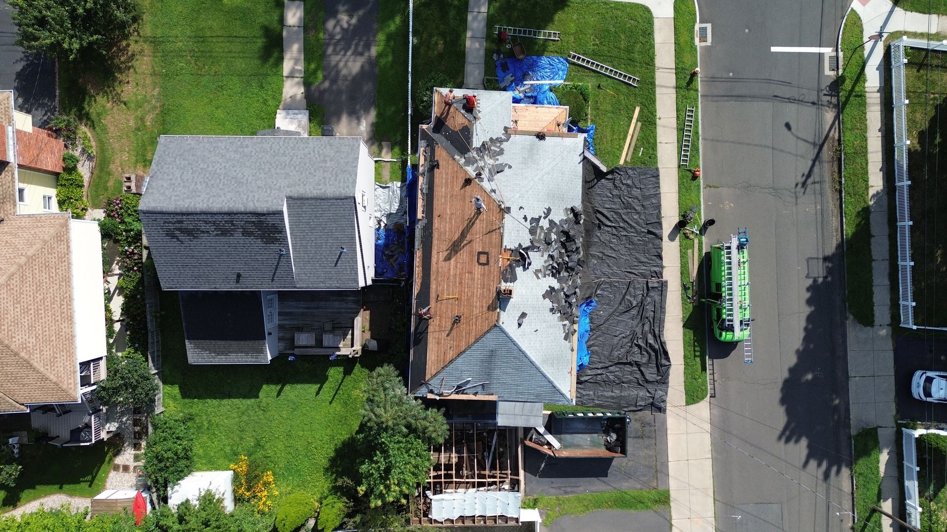 An aerial view of a residential roof replacement in progress, with workers, construction materials, and a dumpster.