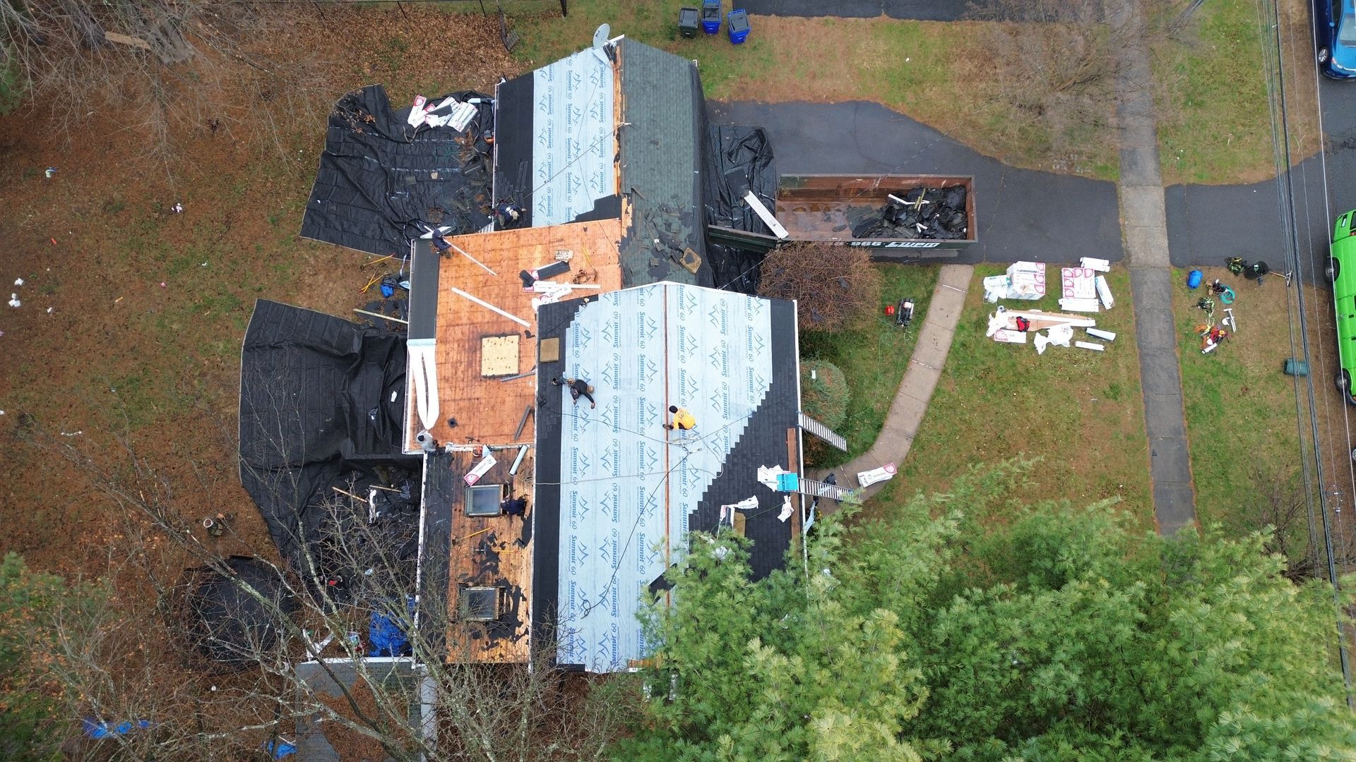 An aerial view shows workers replacing a residential roof, with sections of plywood, underlayment, and roofing materials.