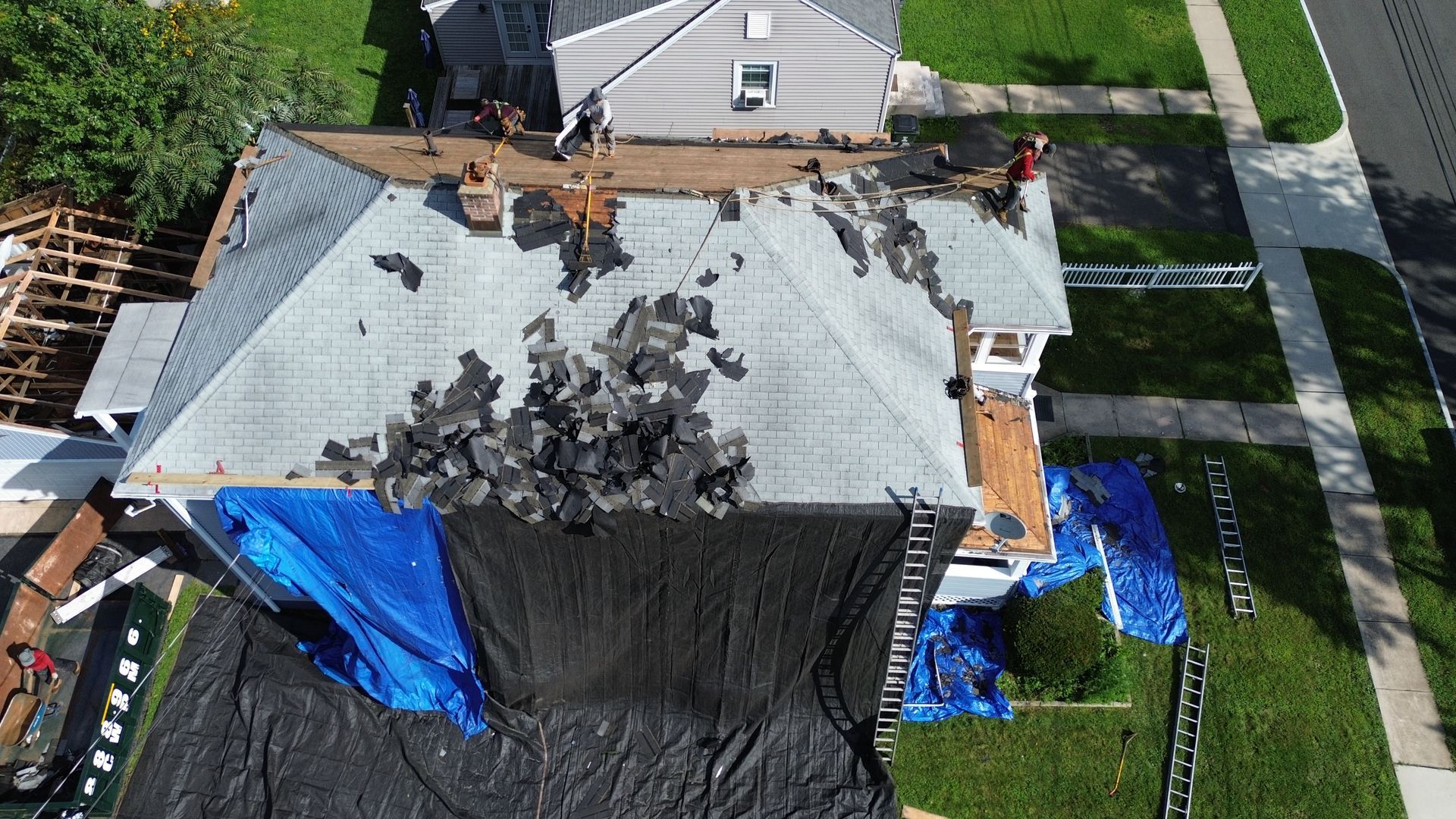 An aerial view of a house roof undergoing repairs, with workers, debris, and protective tarps visible.