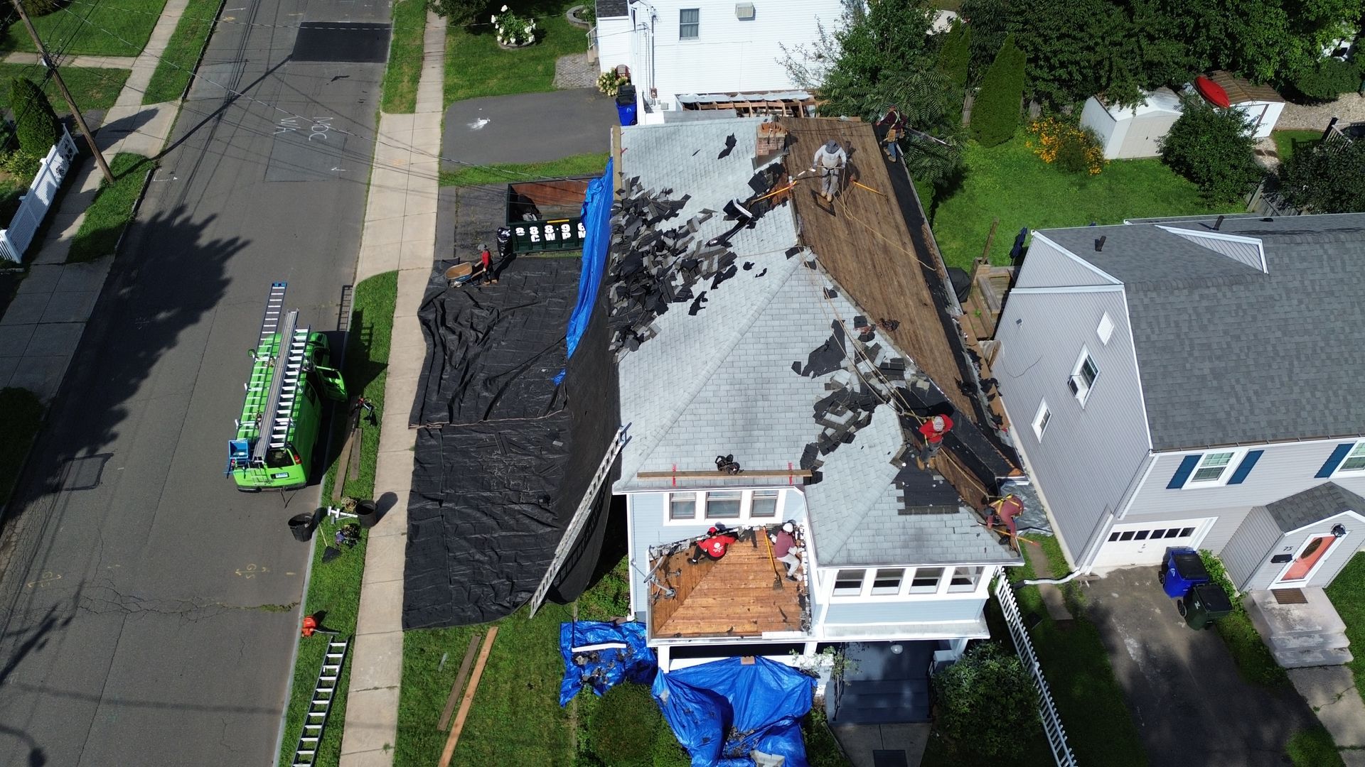 Aerial view of a house undergoing a roof replacement, with tarps covering the yard and a green truck parked nearby.
