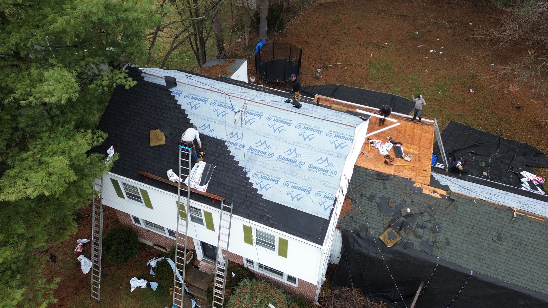 An aerial view of workers installing new black shingles on a residential roof over a light-colored underlayment.