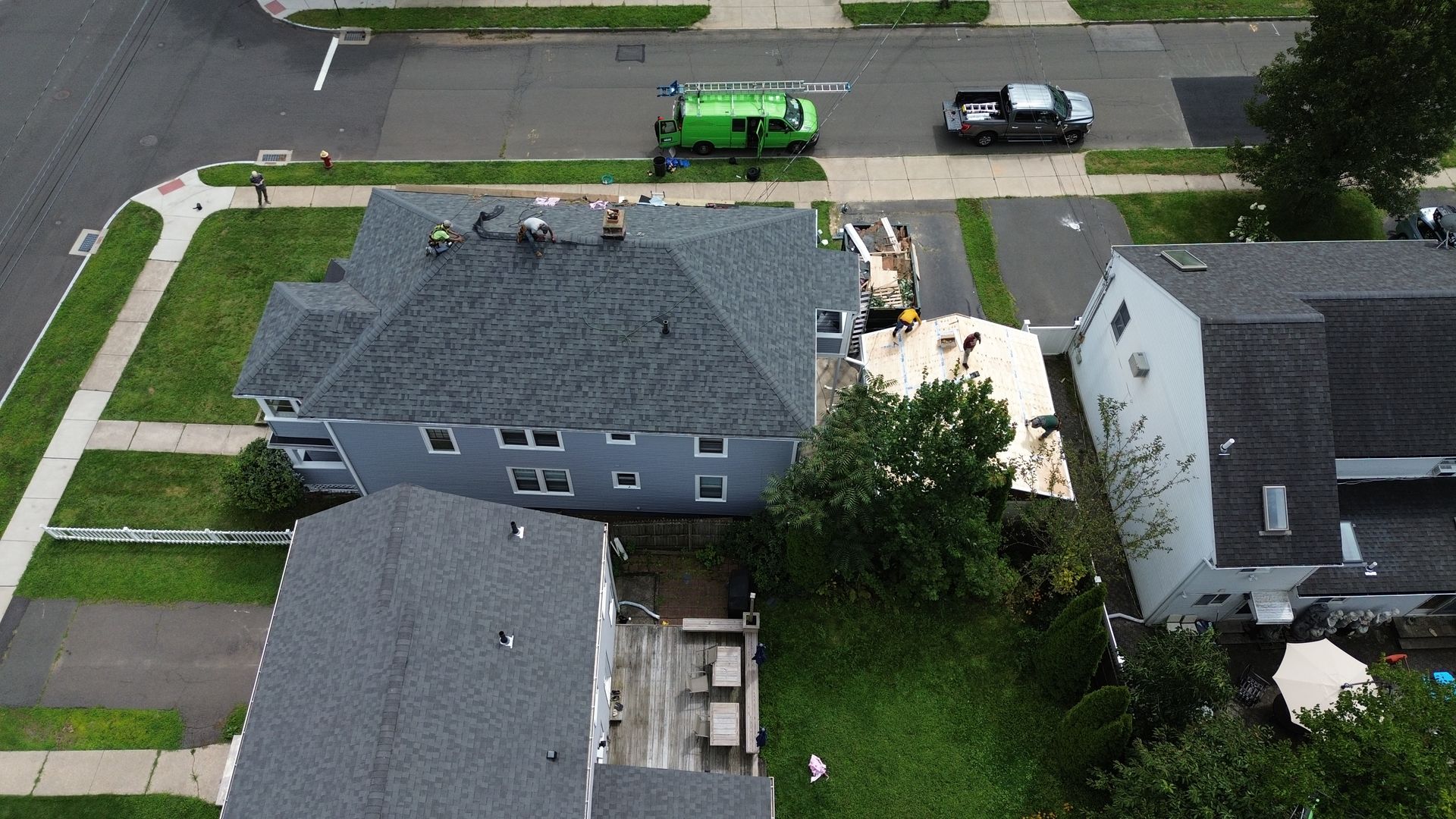 An aerial view shows workers on the roof of a blue house, with construction materials visible near the back entrance.