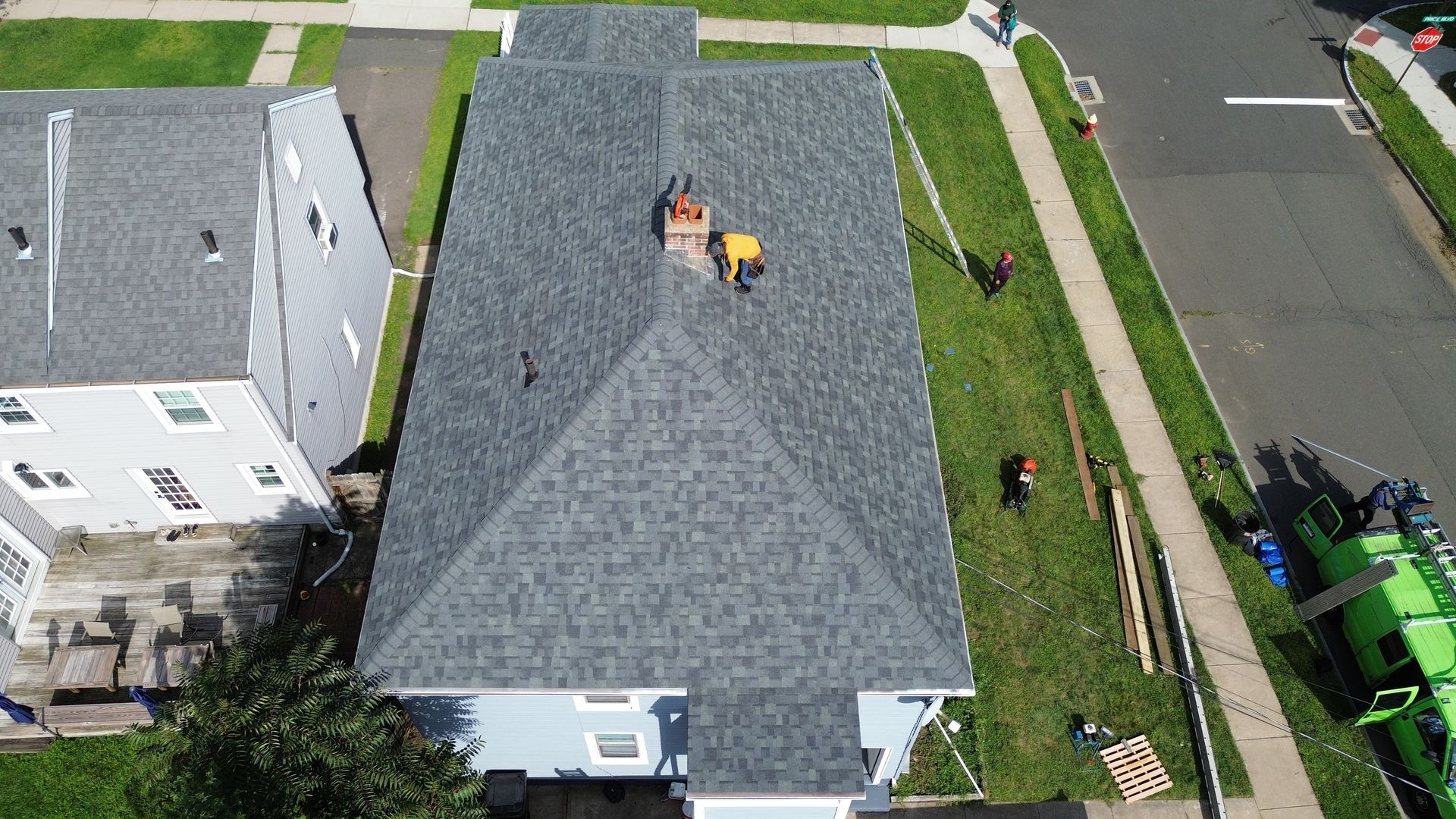 An aerial view shows workers installing dark shingles on a residential roof with a chimney, alongside a parked truck.