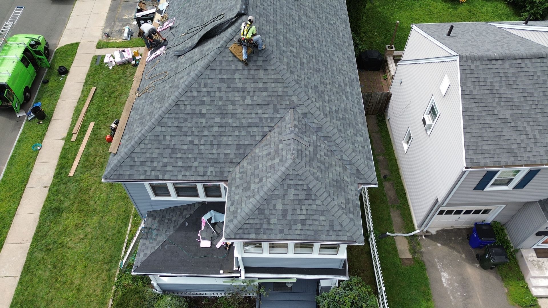 Aerial view of a gray house undergoing roof repairs with workers visible on the shingled surface.
