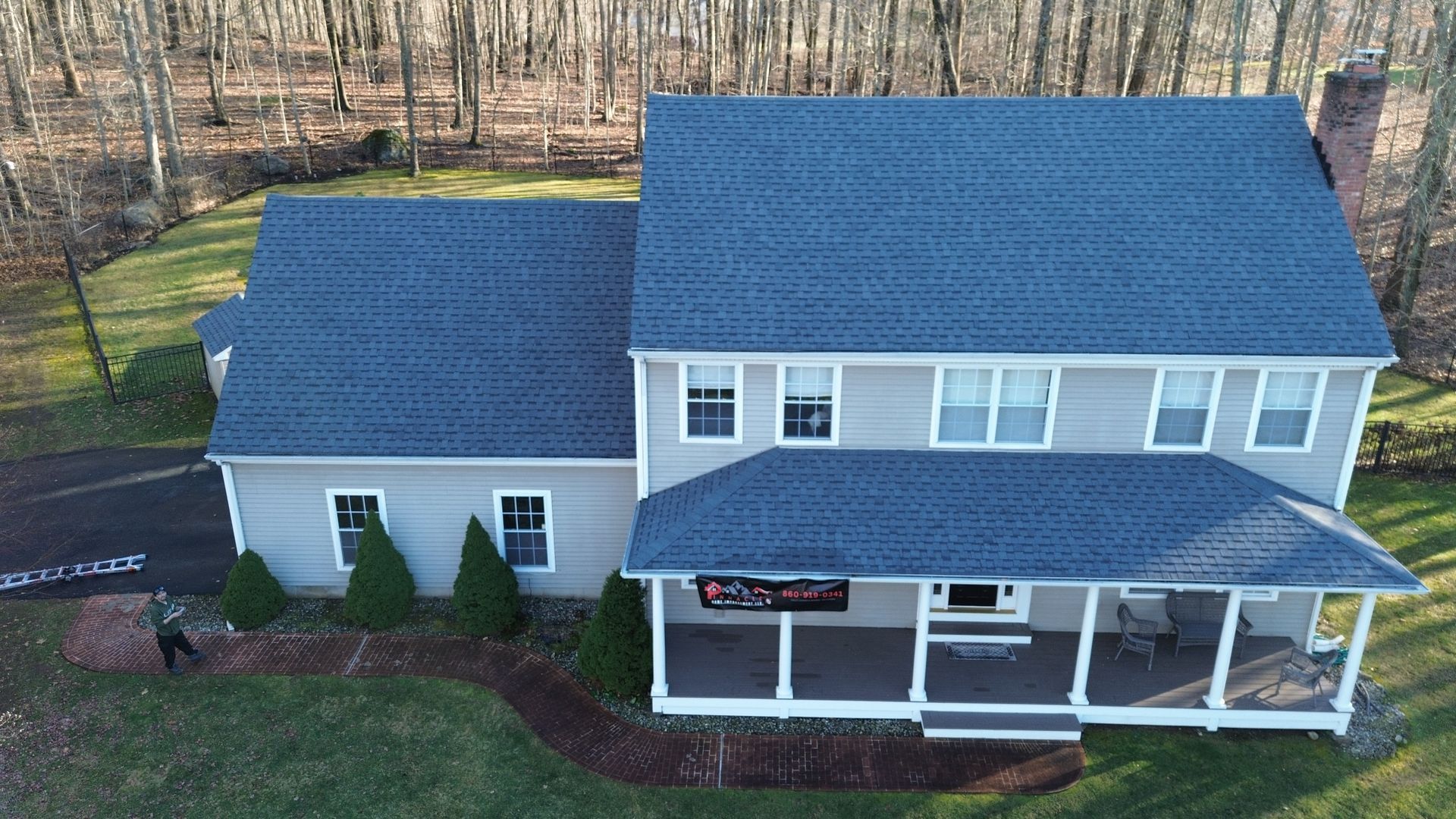 An aerial view of a light-gray two-story house with a blue roof, a front porch, and a winding brick path in a wooded yard.