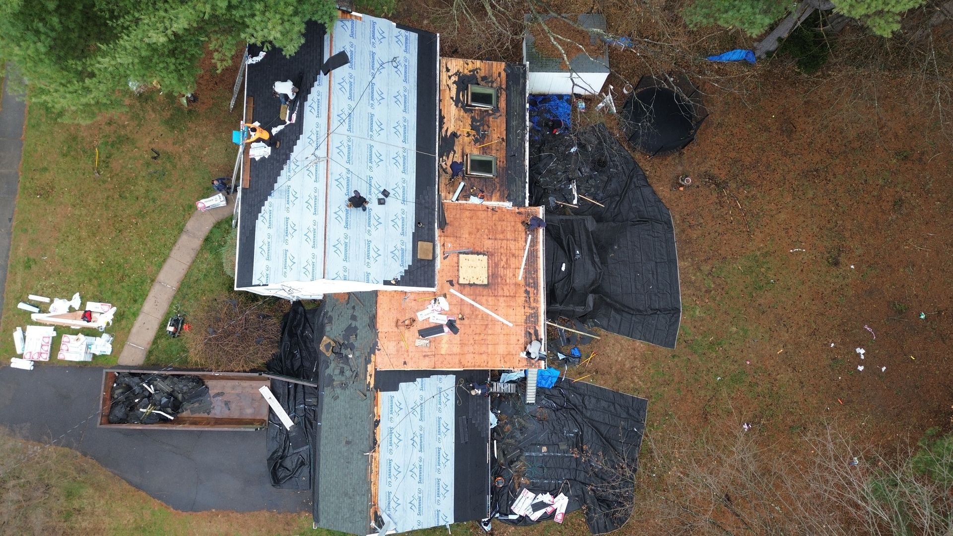 Aerial view of a house undergoing roof replacement with workers, exposed wood, protective underlayment, and a dumpster.