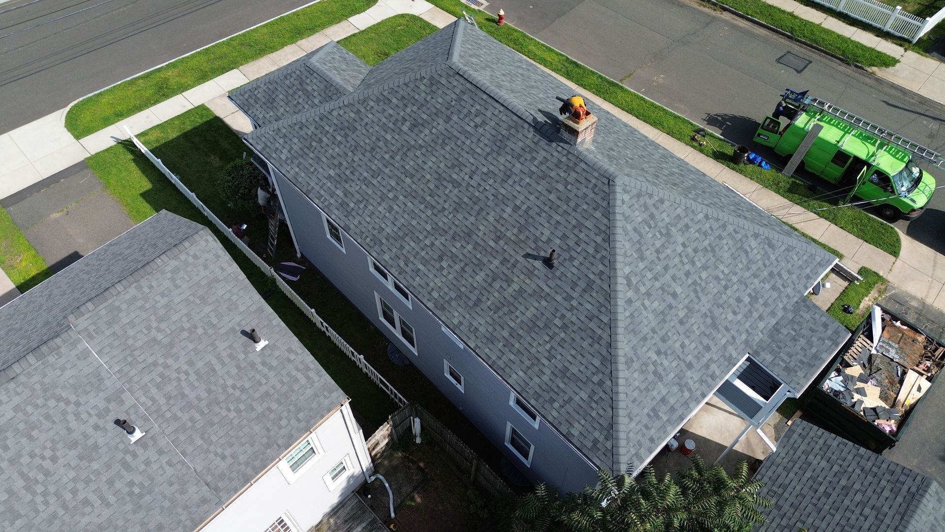 An aerial view of a gray residential house with a new asphalt shingle roof, a chimney, and a construction dumpster nearby.